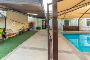 Área de piscina ao ar livre com deck de concreto, grade de metal e área de estar coberta com banco e vasos de plantas.