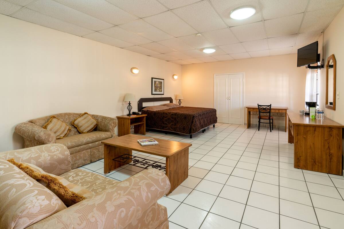 A hotel room with a bed, two sofas, a wooden coffee table, a desk with a chair, and a TV mounted on the wall.