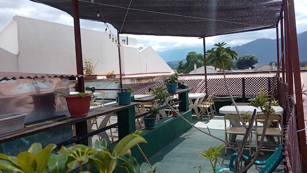 A covered outdoor seating area with tables and chairs, potted plants, and a view of buildings and mountains in the background. - Feathers Hotel and Lounge - Hostel (Antigua Guatemala)