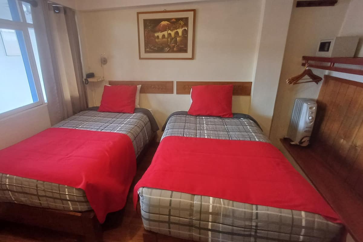 Una habitación con dos camas, cada una con una colcha roja y un edredón a cuadros. Encima de las camas, hay un cuadro enmarcado en la pared, un radiador a la derecha y una ventana a la izquierda.