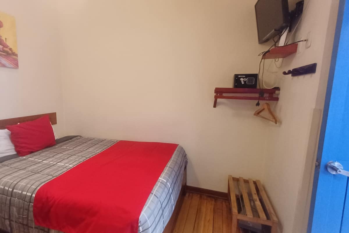 Un dormitorio con una cama, una manta roja, un cabecero de madera, un almohadón blanco y otro rojo, un televisor montado en la pared, un estante pequeño con una radio y un banco de madera.