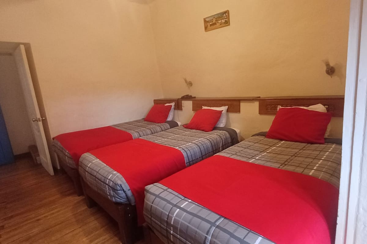 Una habitación con tres camas, cada una con una manta roja y una colcha a cuadros. Se observa una puerta a la izquierda y un cuadro colgado en la pared sobre las camas.