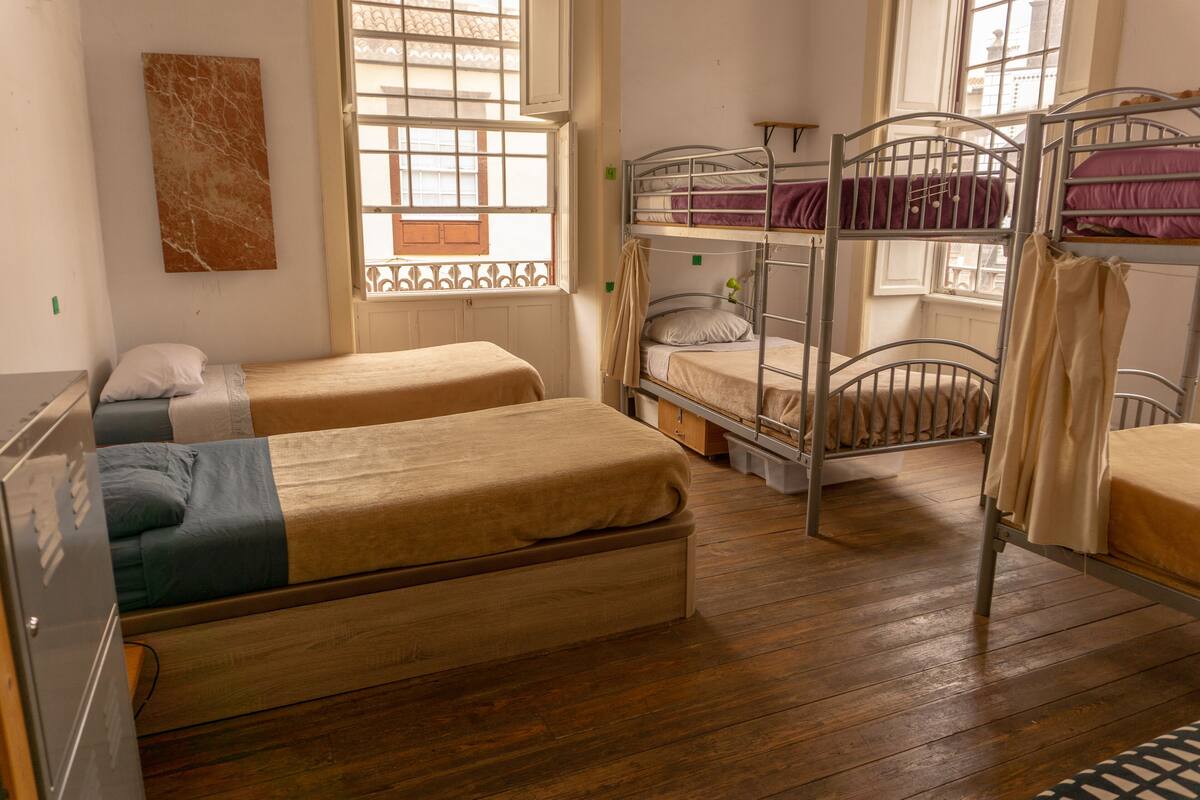 A dormitory room with bunk beds, wooden flooring, a window with bars, and a wall-mounted artwork.