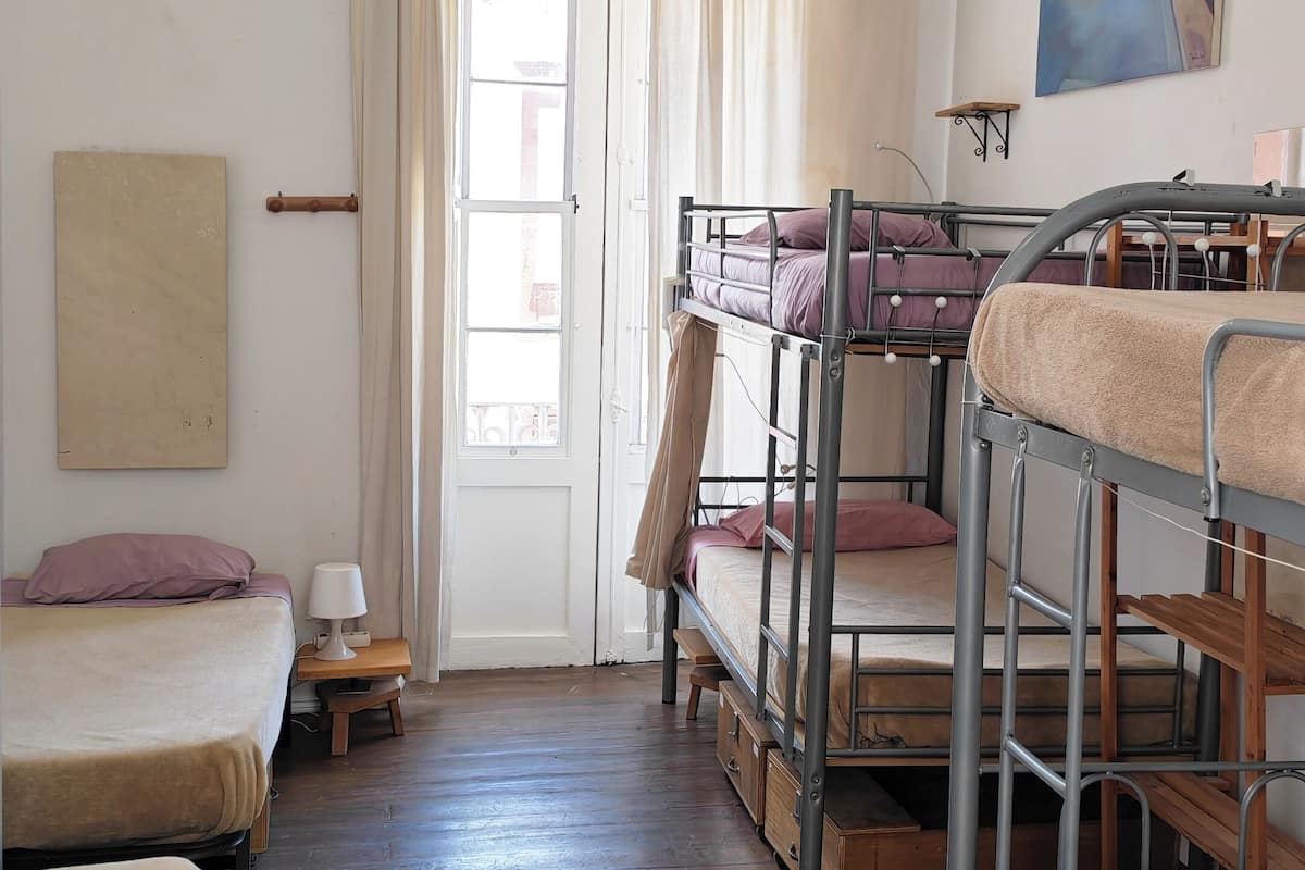 A dormitory room with bunk beds, a hanging light fixture, a painting on the wall, and a window with curtains.