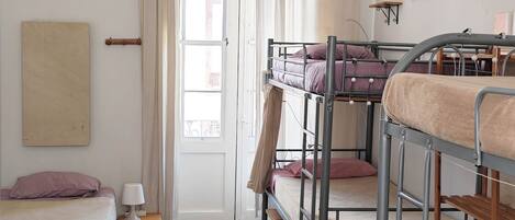 A dormitory room with bunk beds, a hanging light fixture, a painting on the wall, and a window with curtains.