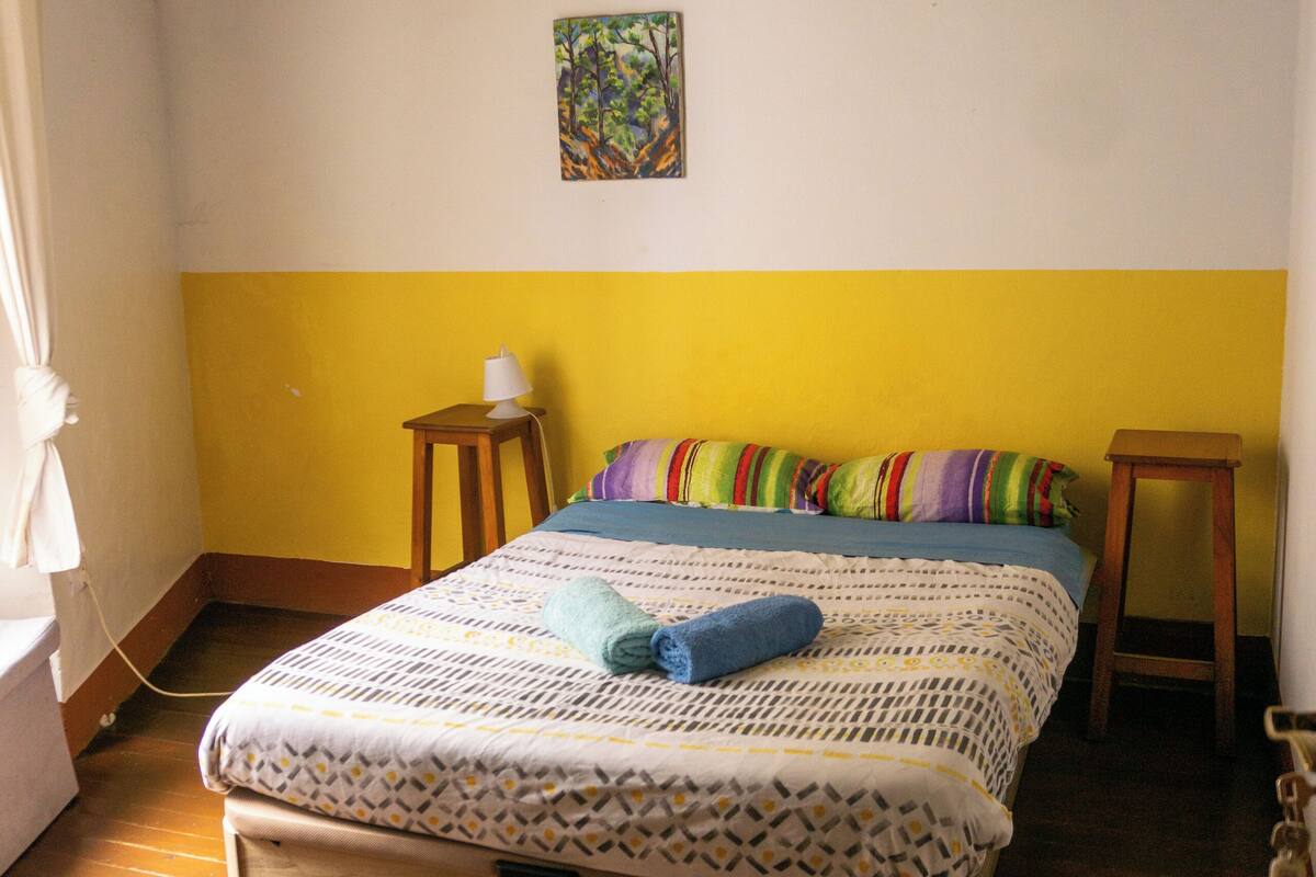A bedroom with a bed, a bedside table, a hanging light, and a wall painting.