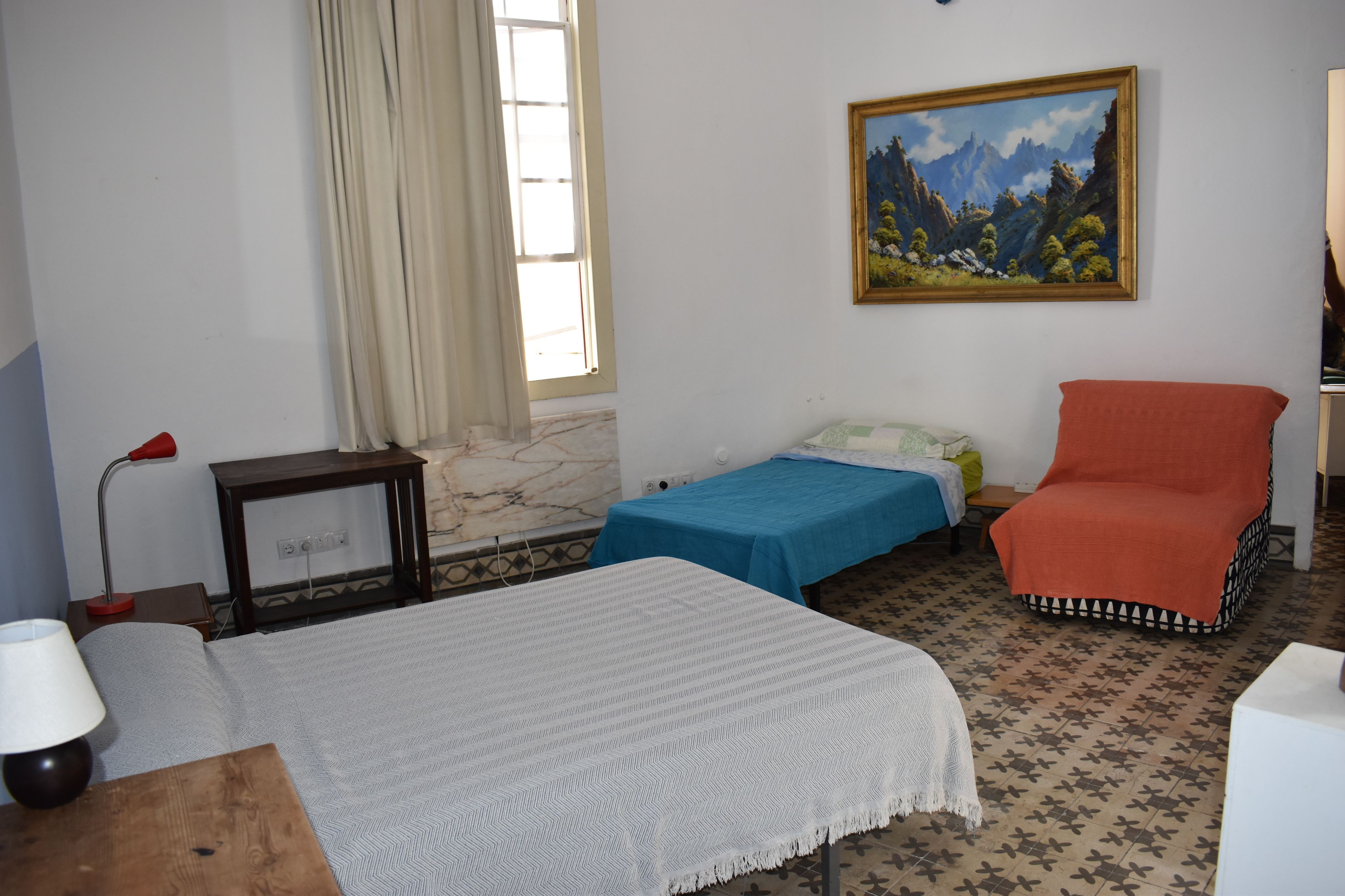 Una habitación con una cama, una silla, una mesa, una lámpara, un cuadro y una ventana con cortinas.