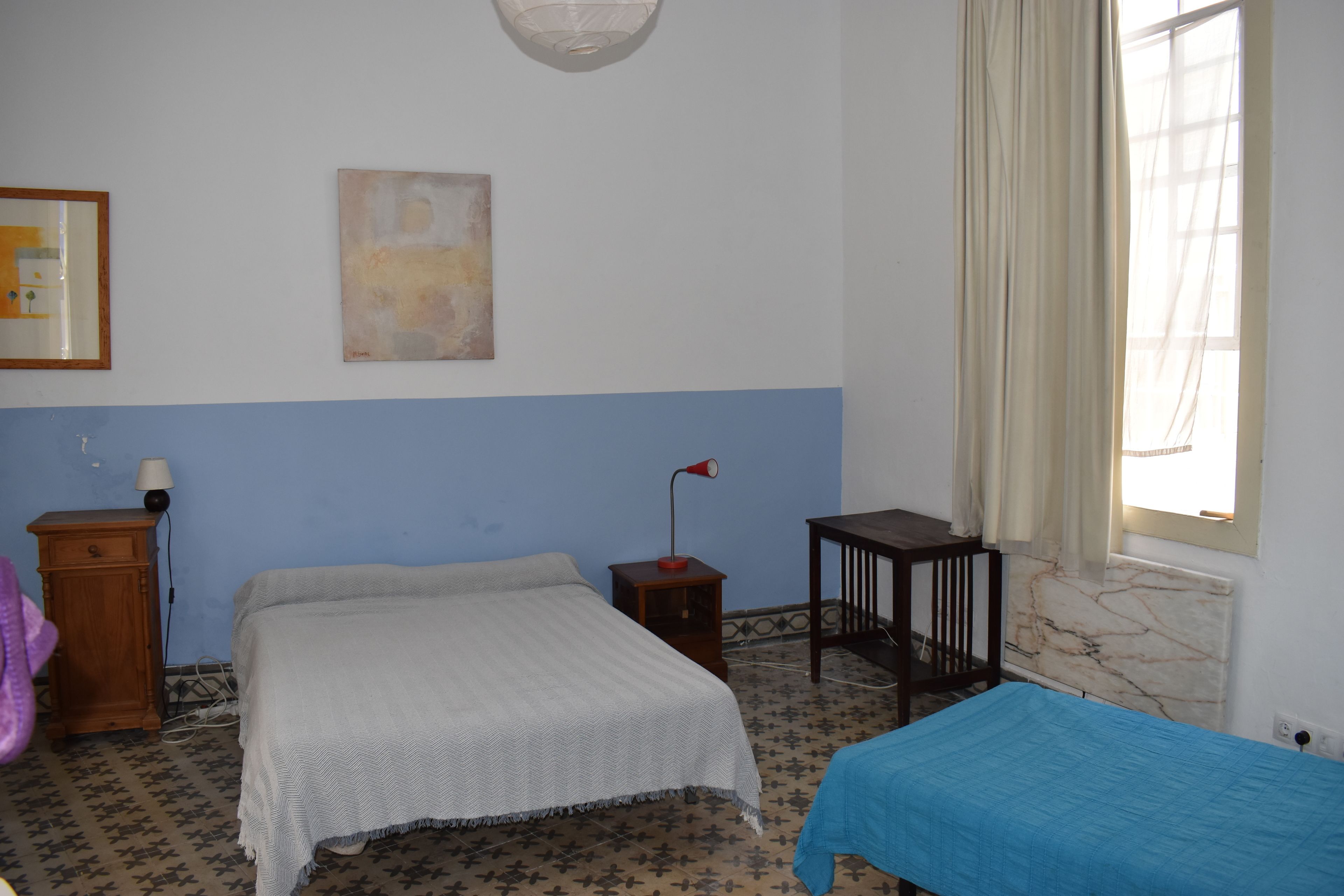A bedroom with a bed, a small table, a nightstand, a lamp, and a window with curtains.
