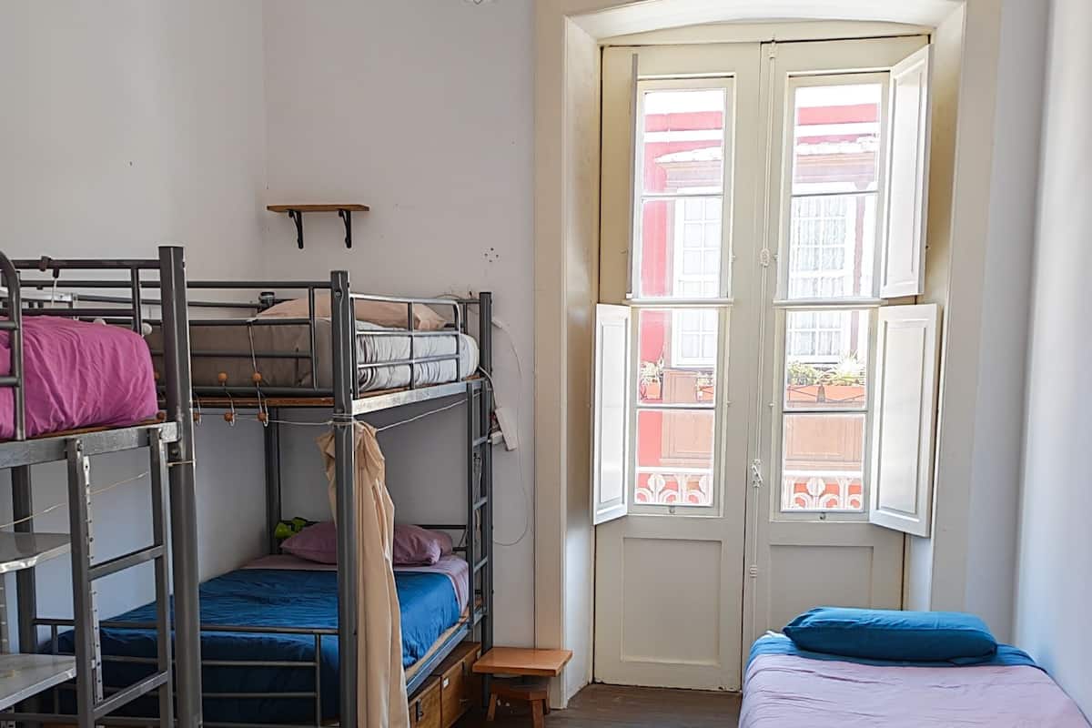 A dormitory room with bunk beds, a small table, and a window with a view of buildings.