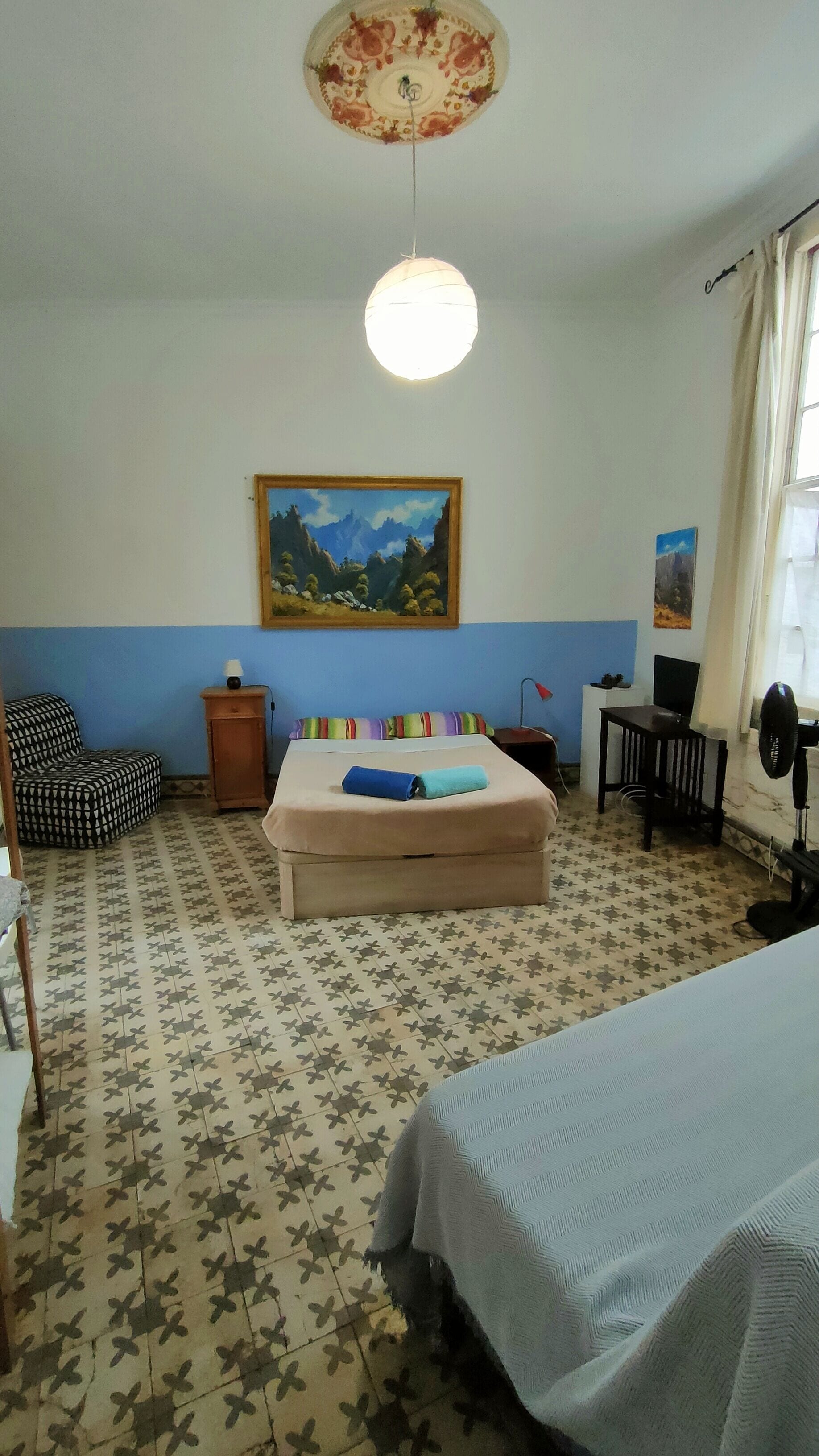 A hotel room with a bed, a chair, a desk, a lamp, and a painting on the wall.