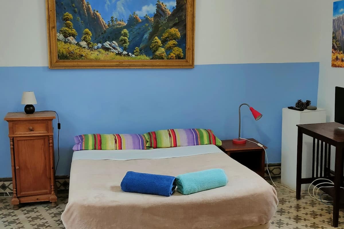A bedroom with a bed, a nightstand, a desk, a chair, a painting, and a lamp.