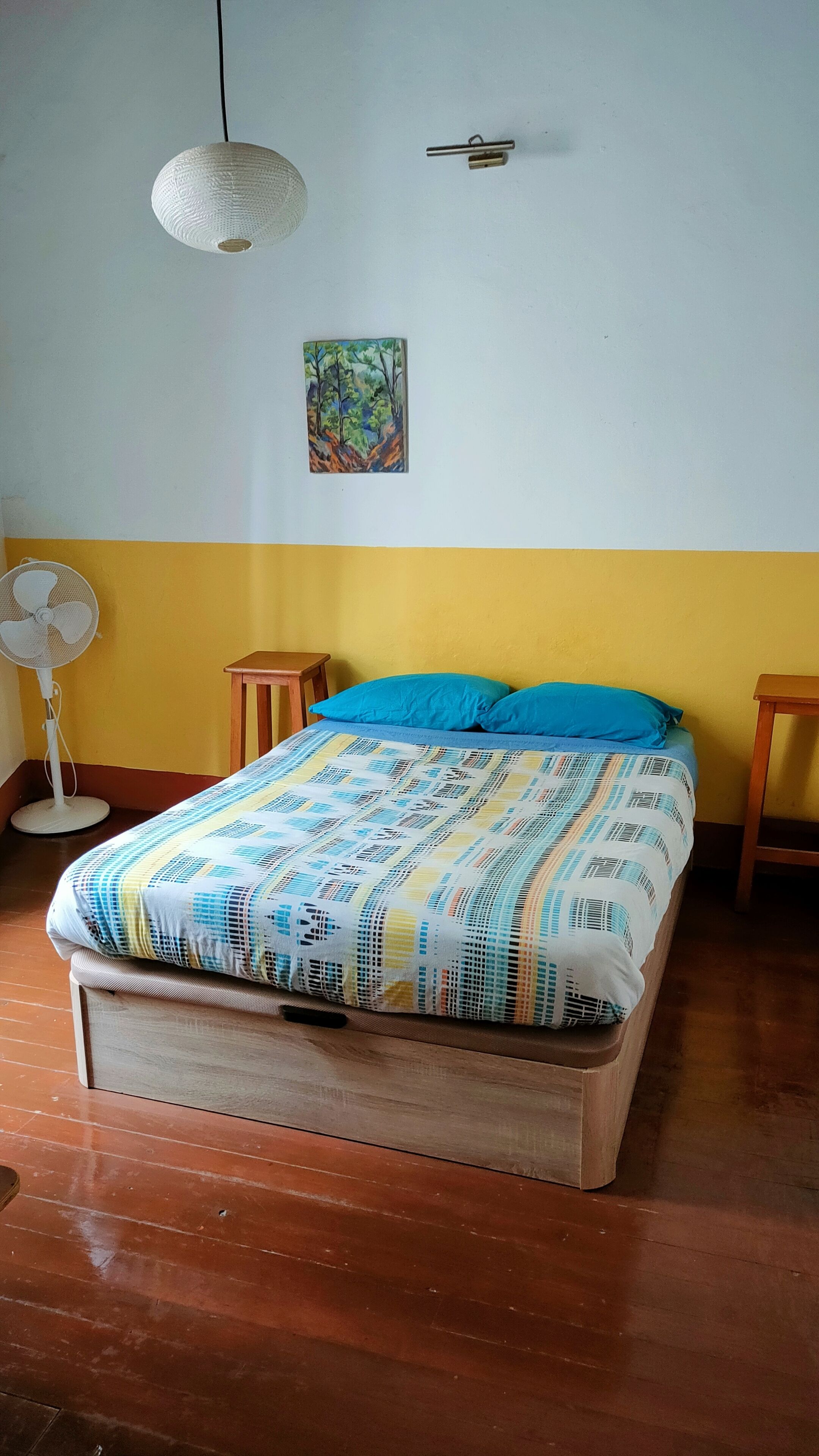 Una camera da letto con un letto, una ventola a parete, un quadro e un comodino in legno.