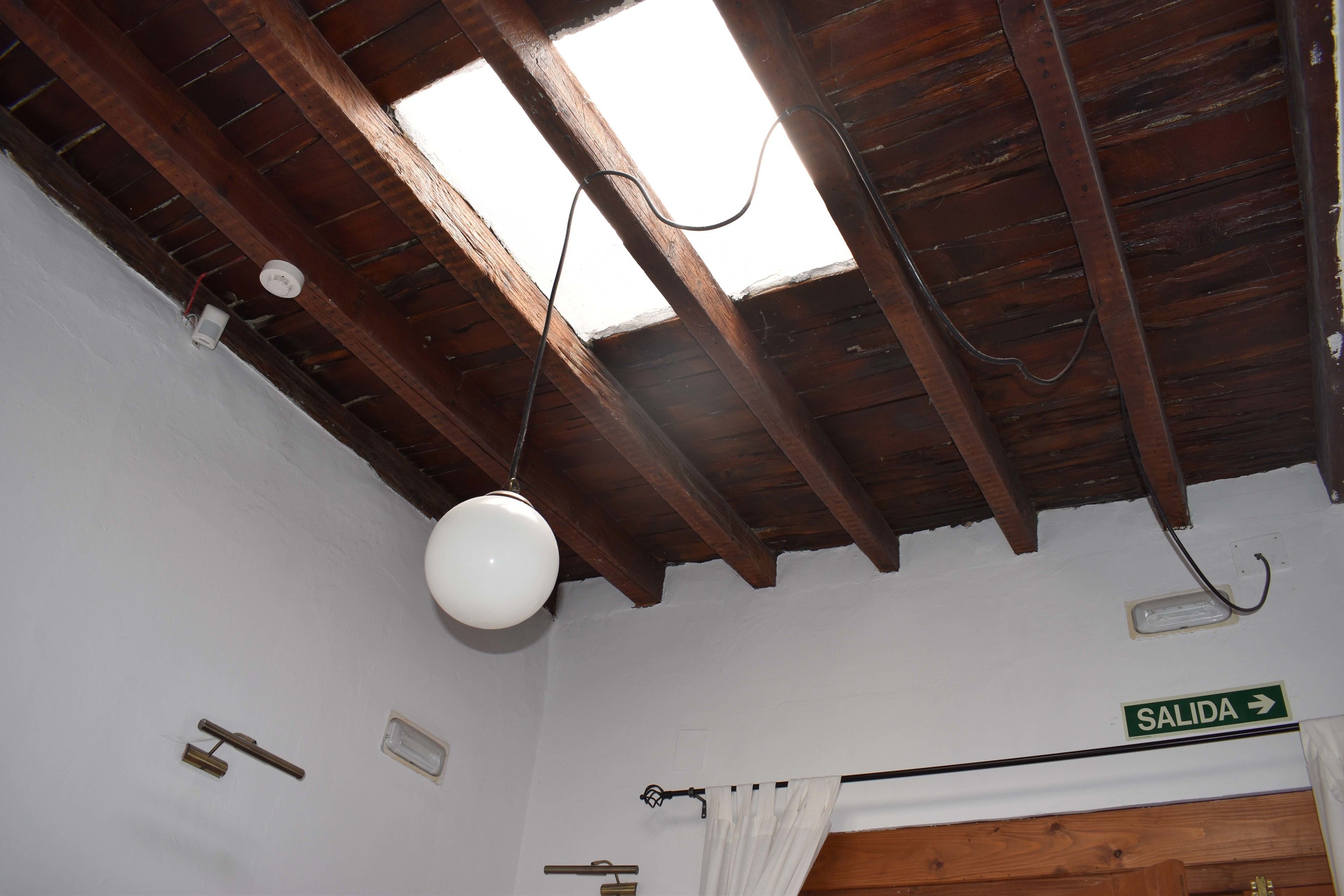 Una habitación rústica con vigas de madera a la vista, tragaluz, ventilador de techo blanco, un cartel que indica una salida y un perchero.