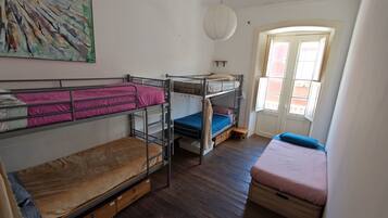 A dormitory room with bunk beds, a wooden floor, a window with curtains, and a hanging light fixture.