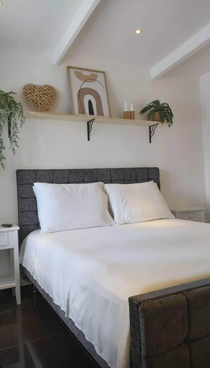 A neatly made bed with white linens, a dark headboard, and a shelf above with decorative items and plants. - KDF Apartments (Willemstad)