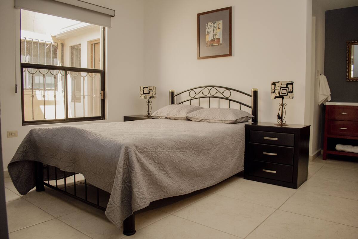 Un dormitorio con cama, mesitas de noche, cómoda, ventana con vistas y un cuadro en la pared.