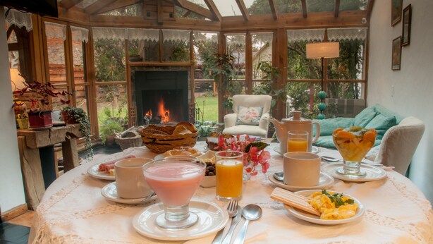 A breakfast table set with various dishes, drinks, and a fireplace in the background. - Hosteria El Arbol Duende (San Martín de los Andes)