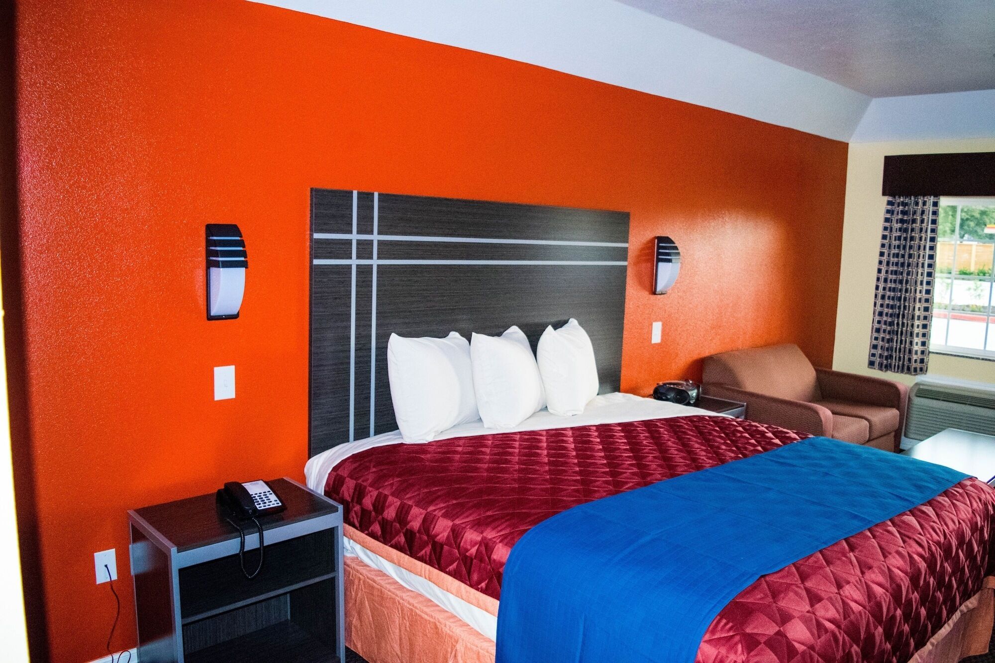 A hotel room with a large bed, a nightstand with a phone, a chair, and a window with curtains.