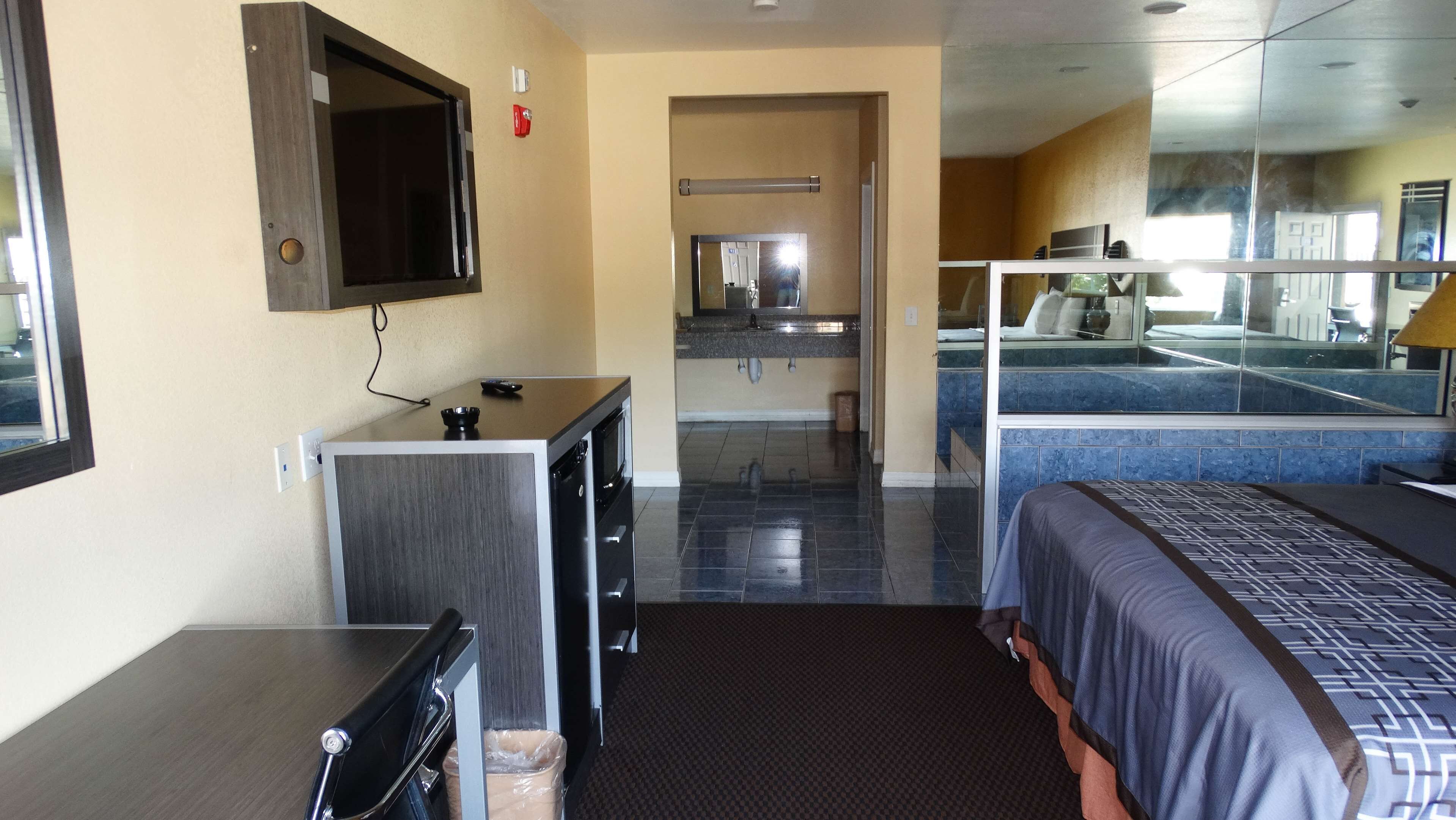A hotel room with a bed, a desk with a chair, a small kitchenette, and a bathroom with a mirror.