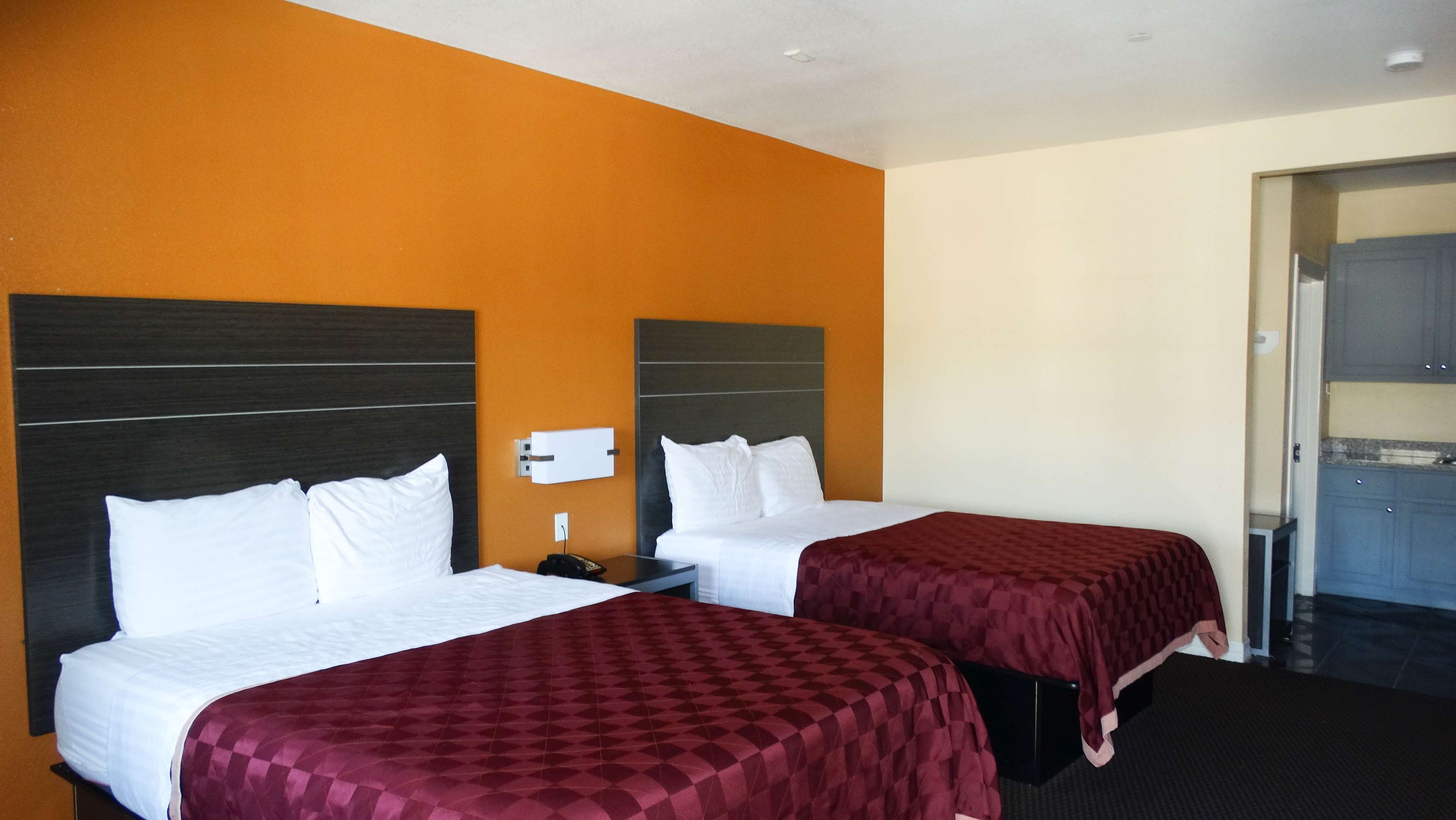 Ett hotellrum med två sängar, båda med vita sängkläder och en mörk sänggavel. Det finns ett litet nattduksbord med en lampa och en telefon. Rummet har orangefärgade väggar och en dörr som leder till ett litet pentry.