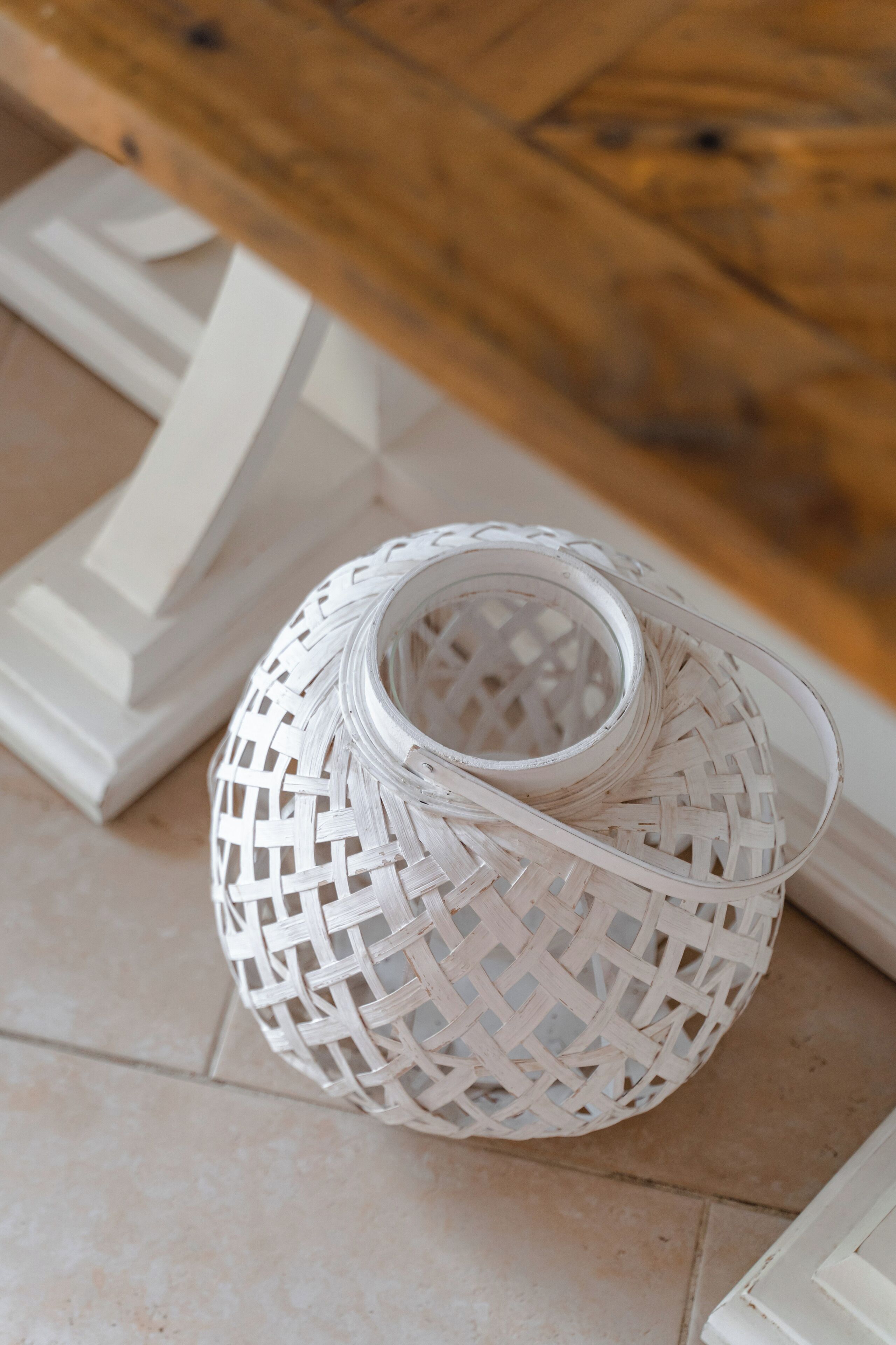 Un farol tejido cilíndrico con un diseño de celosía, sobre un suelo de baldosas cerca de un pilar blanco y una superficie de madera.