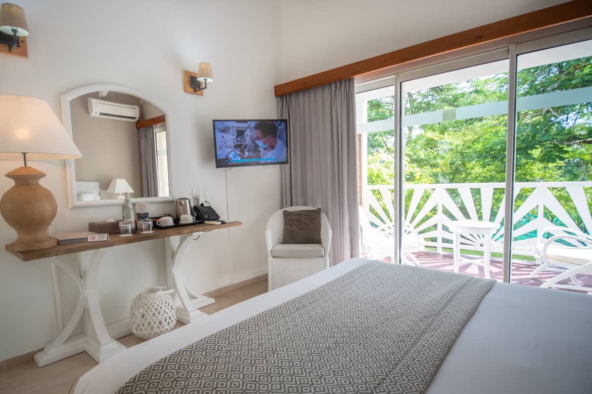 A hotel room with a bed, a desk with a lamp, a television, and a balcony with white chairs.