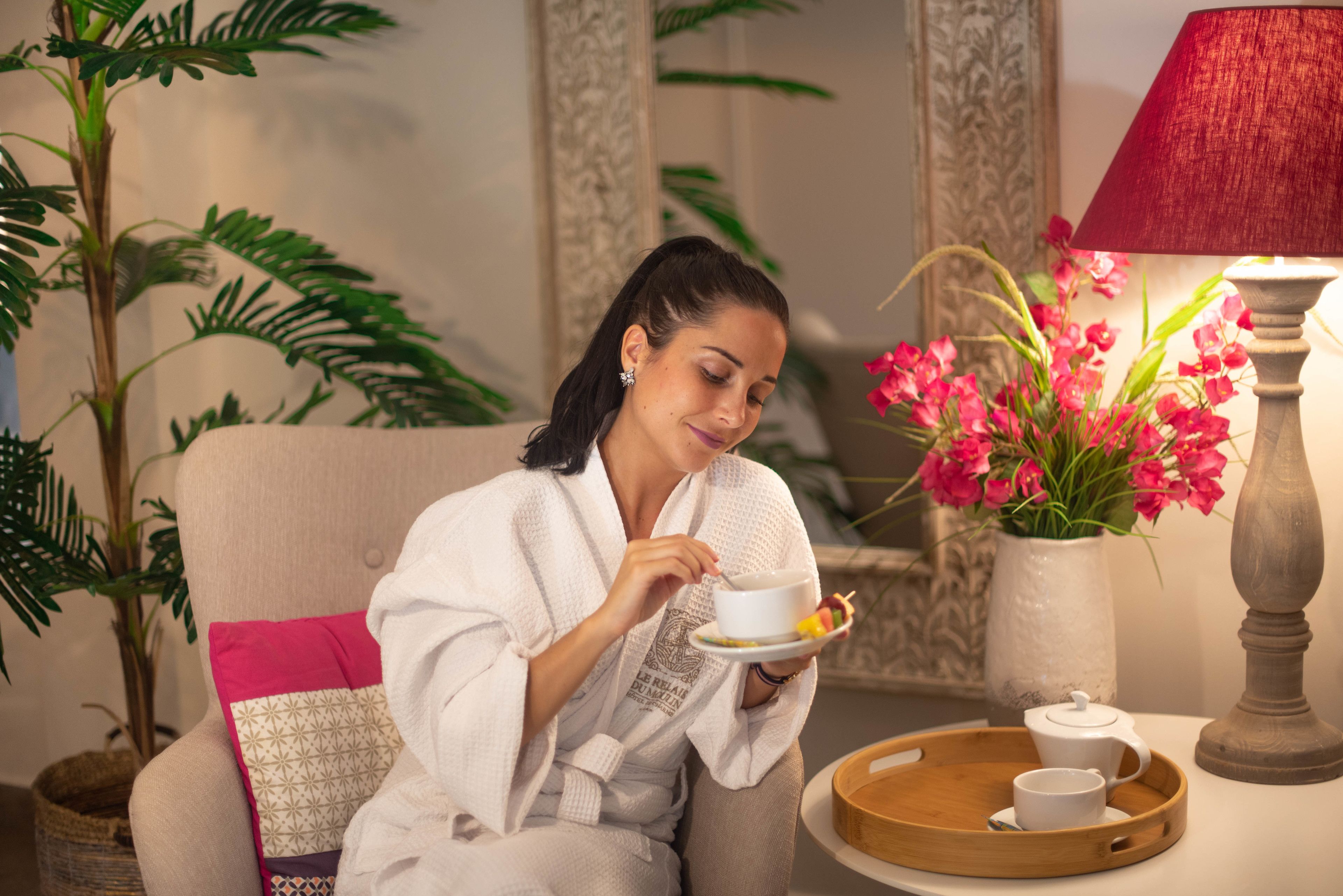 A person in a white bathrobe sitting on a beige sofa with a tray of tea and a teapot.