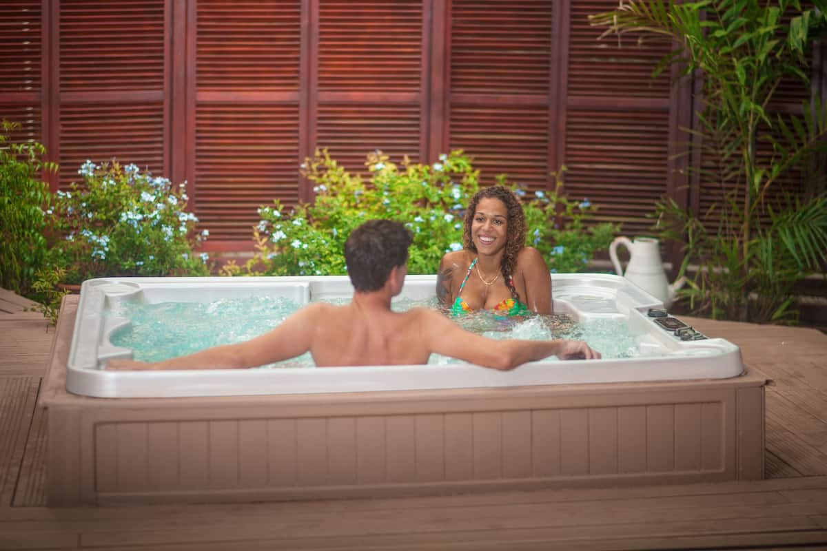 A couple in a hot tub with a wooden deck and greenery in the background.