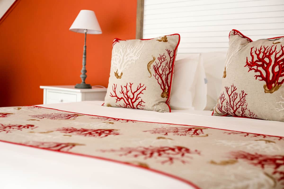 A bed with a beige and red patterned bedspread, two pillows with red coral designs, a white bedside table, and a lamp.