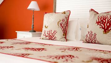 Una cama con colcha estampada en tonos beige y rojo, dos almohadas con diseños de coral rojo, una mesita de noche blanca y una lámpara.