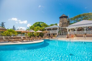 Un área de piscina con sillas de descanso y sombrillas, una torre de piedra y un edificio con techo de paja.