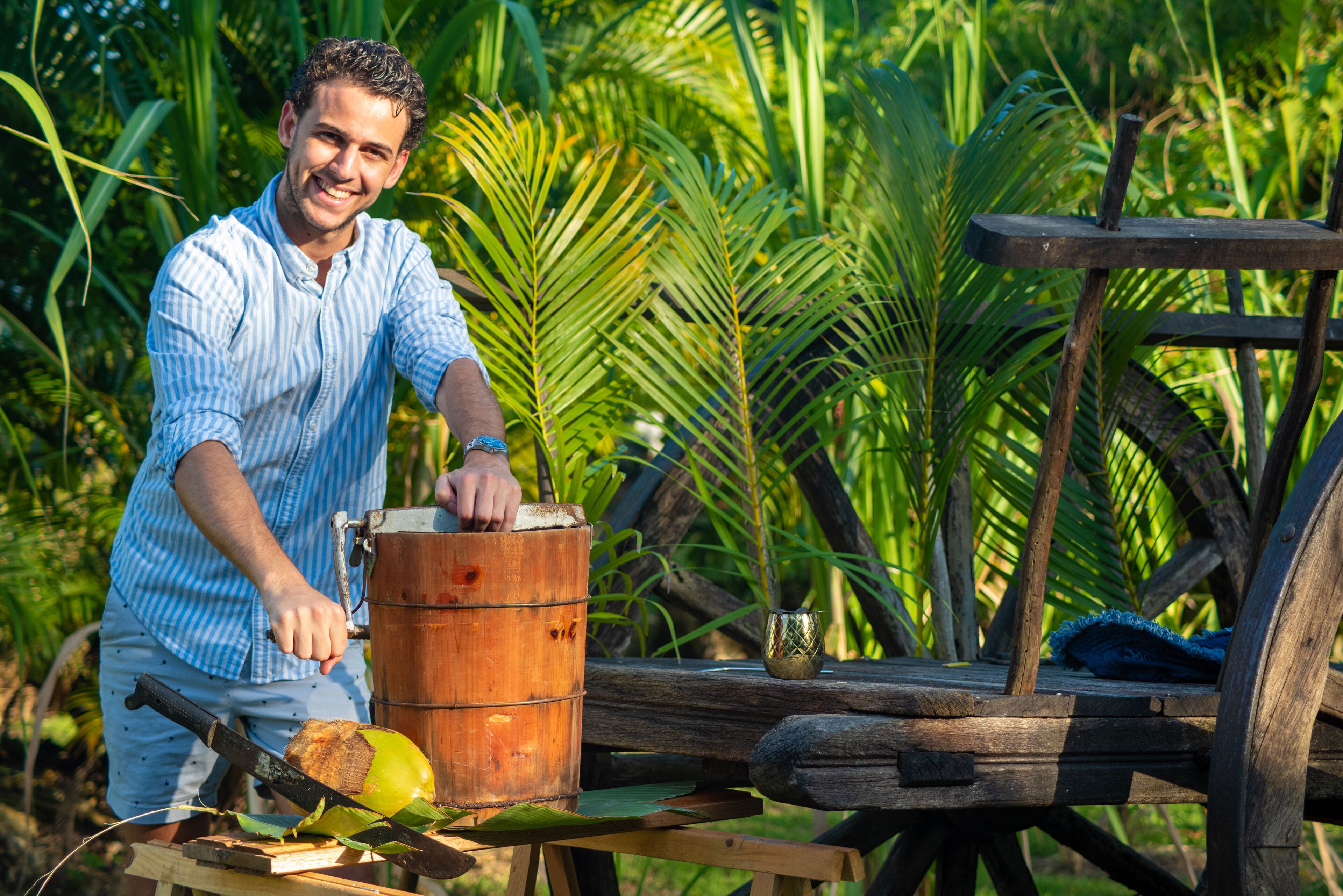 Une personne utilise un extracteur de jus manuel en bois pour presser une noix de coco sur une table en bois, à l'extérieur, dans un environnement luxuriant et verdoyant.