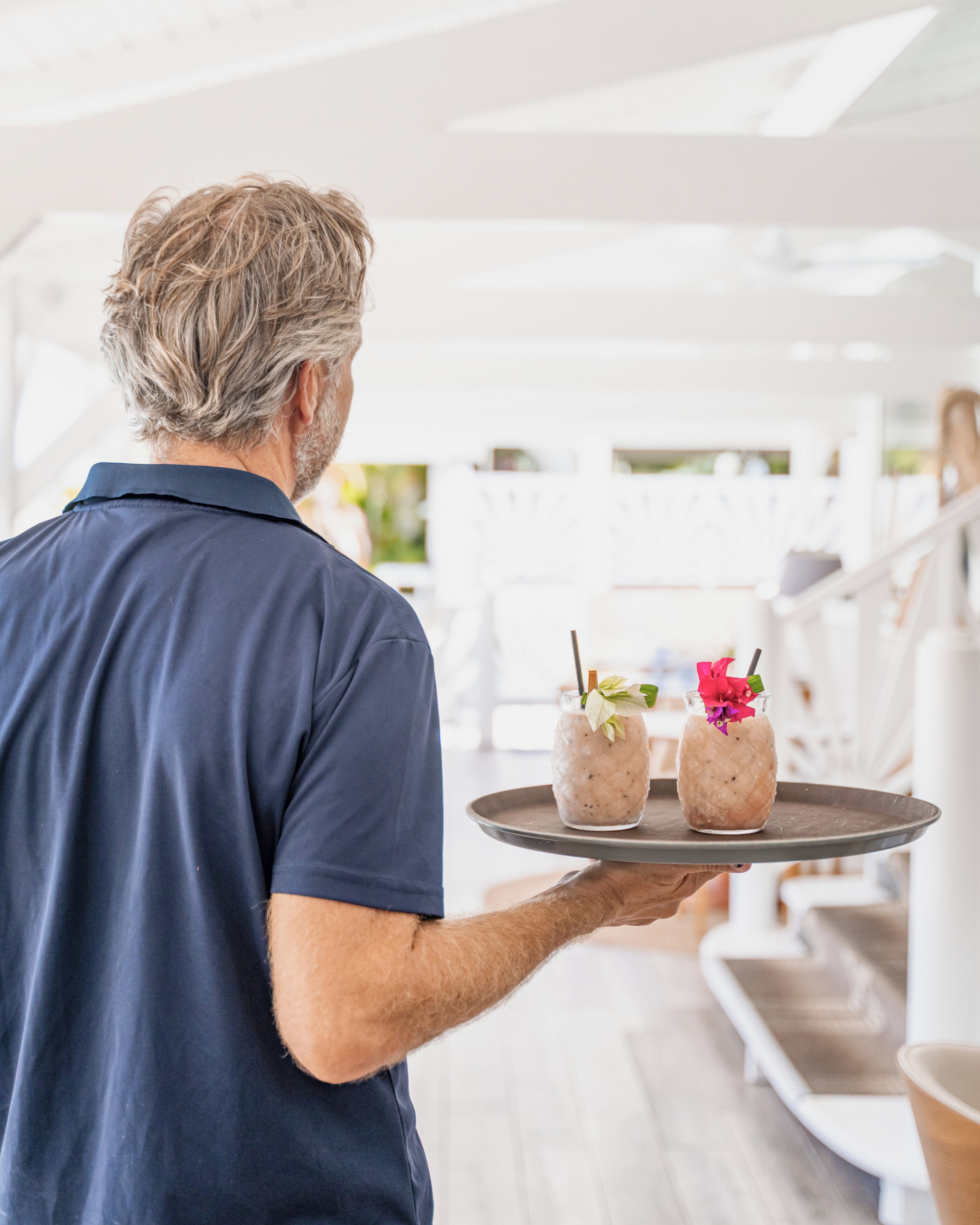 Una persona sosteniendo una bandeja con dos bebidas, una con una flor rosa, en un ambiente interior moderno y luminoso con mobiliario blanco.