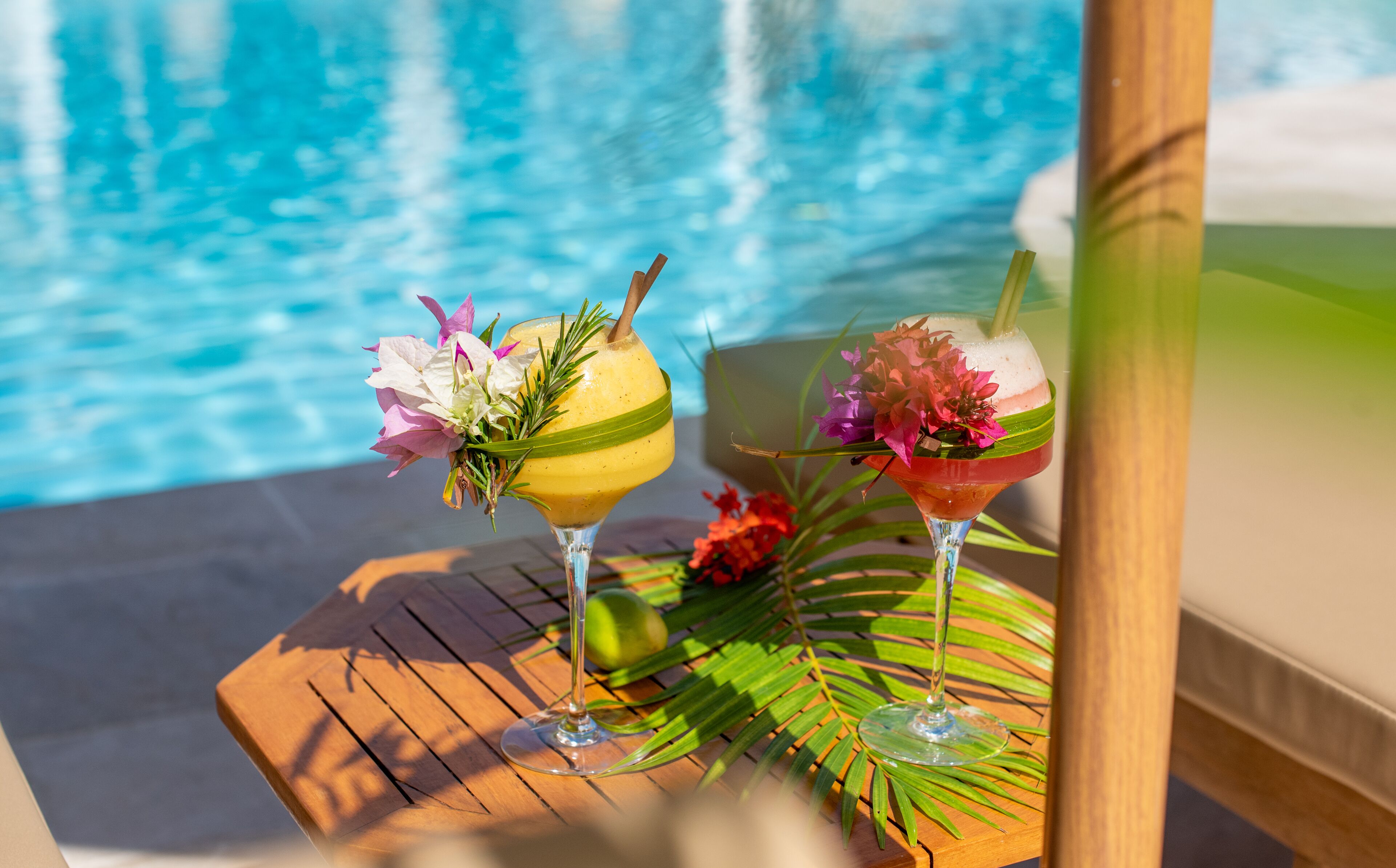 Twee cocktails op een houten tafel bij een zwembad, in een tropische omgeving.