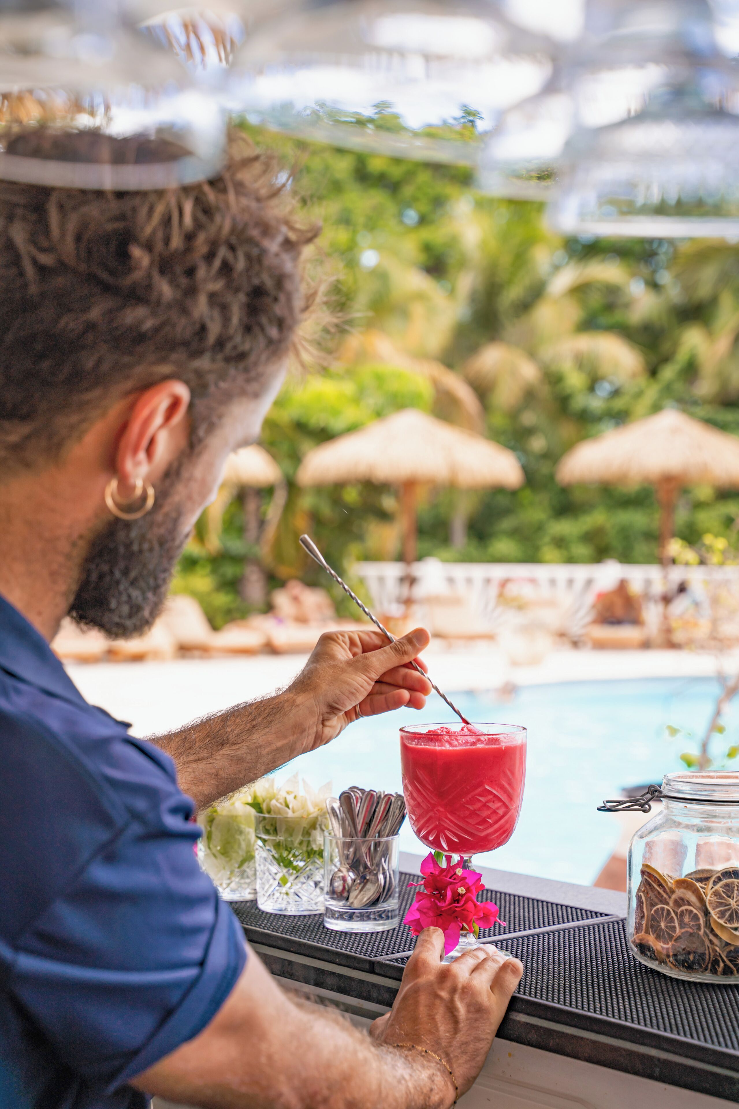 Una persona sentada junto a una mesa al lado de la piscina, con una bebida y un vaso de jugo, rodeada de sombrillas y follaje.