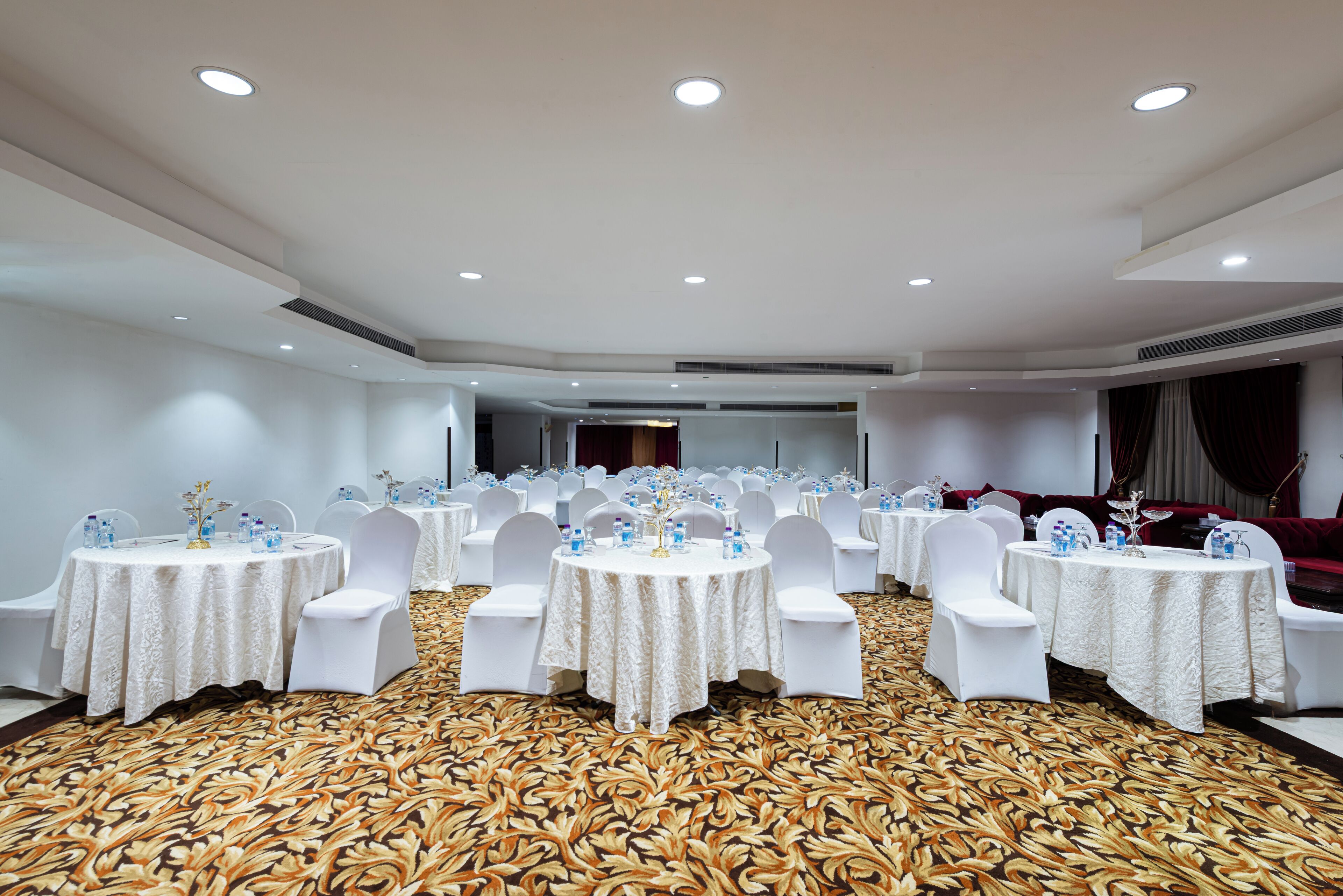 Une salle de banquet avec des tables rondes, des chaises blanches et un tapis à motifs.