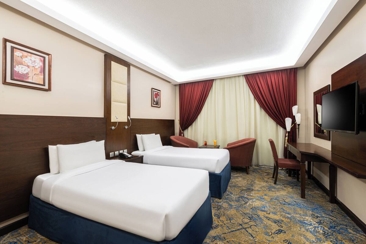A hotel room with two beds, a desk, a chair, a TV, and a large window with curtains.