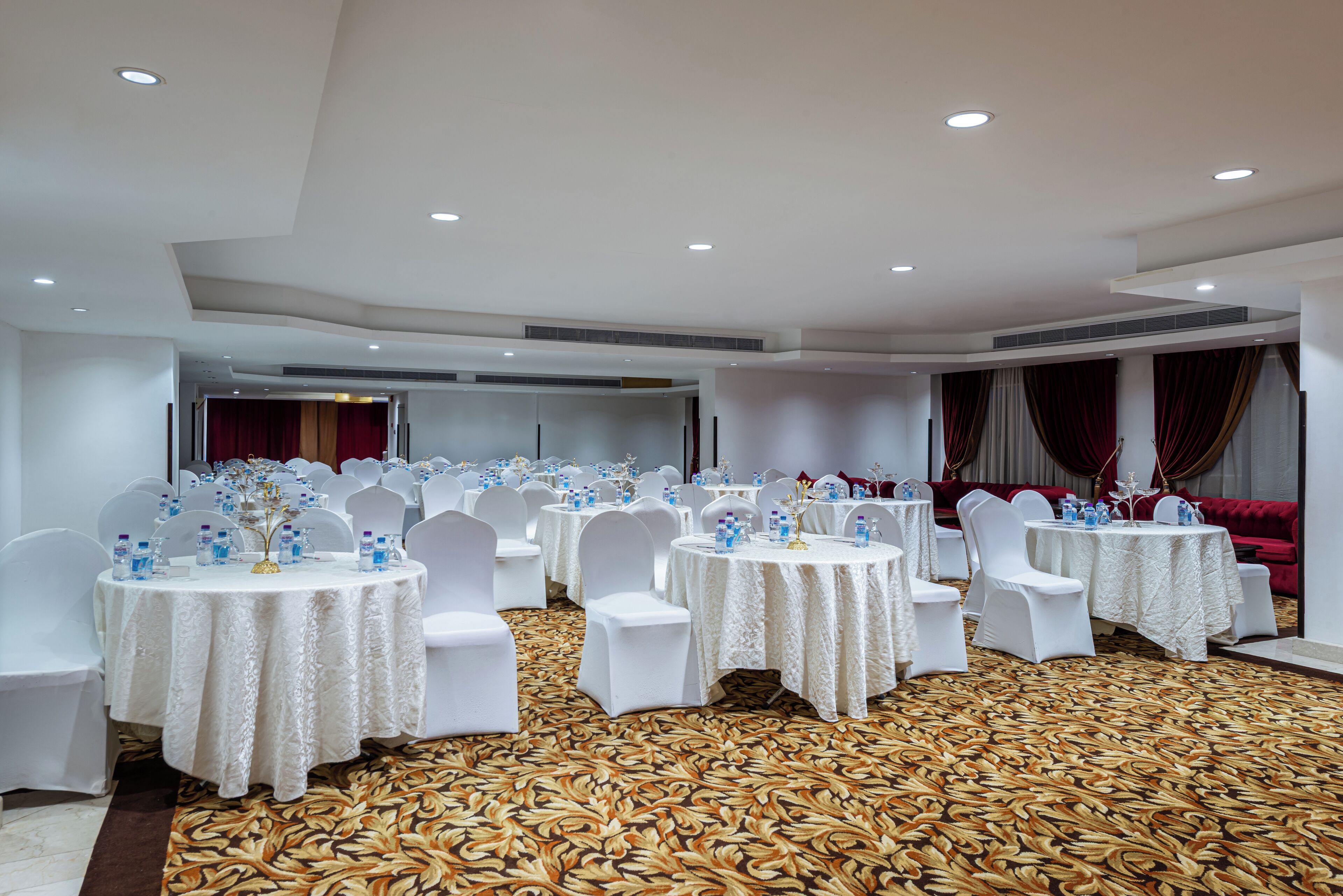 A banquet hall with round tables, white chairs, and a patterned carpet.