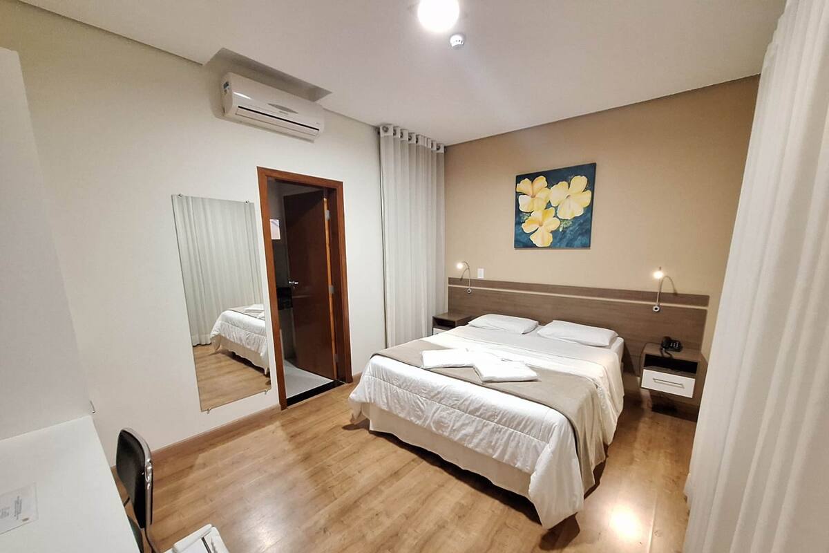 A hotel room with a large bed, a bedside table, a nightstand, a mirror, and an air conditioning unit.