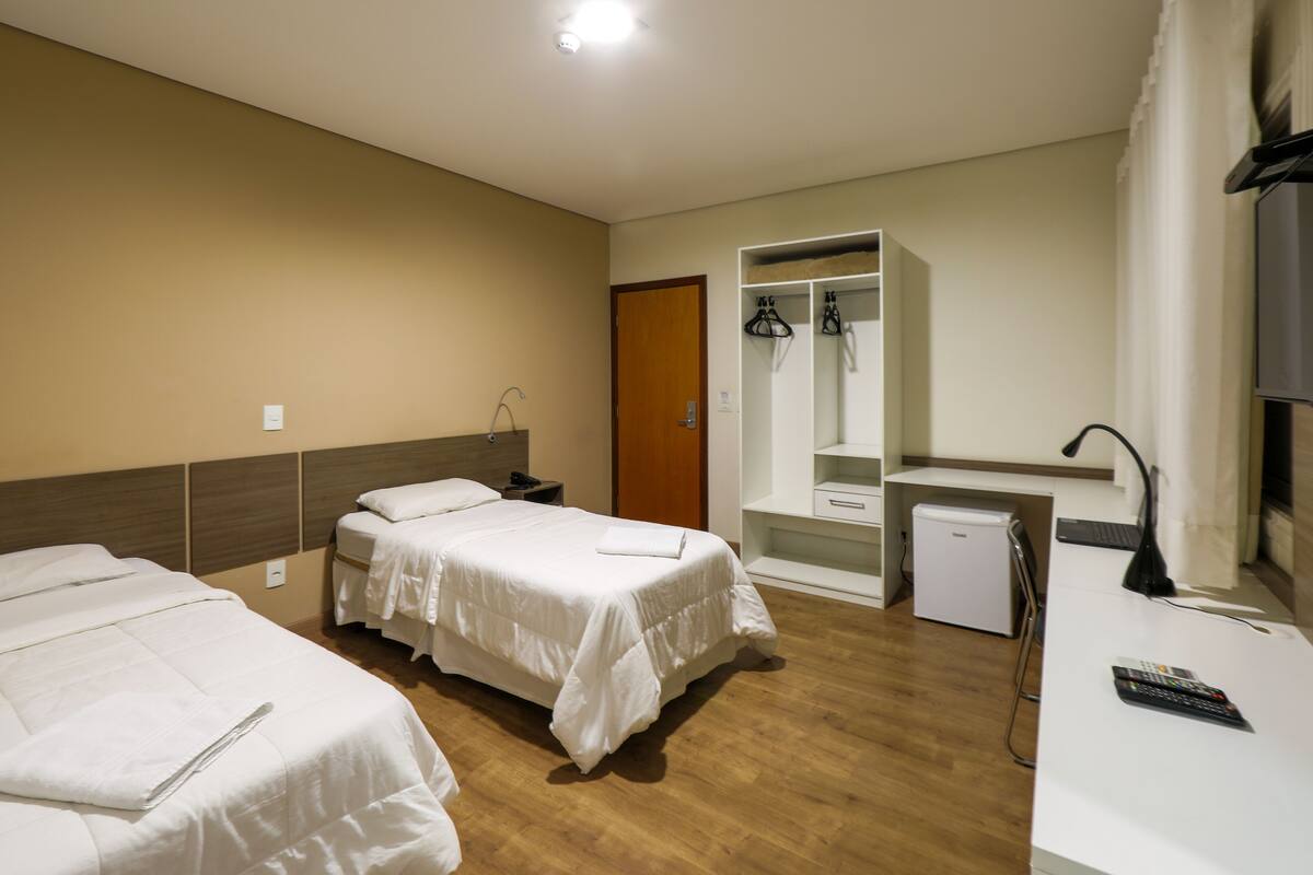 A hotel room with two beds, a desk, a wardrobe, and a small refrigerator.