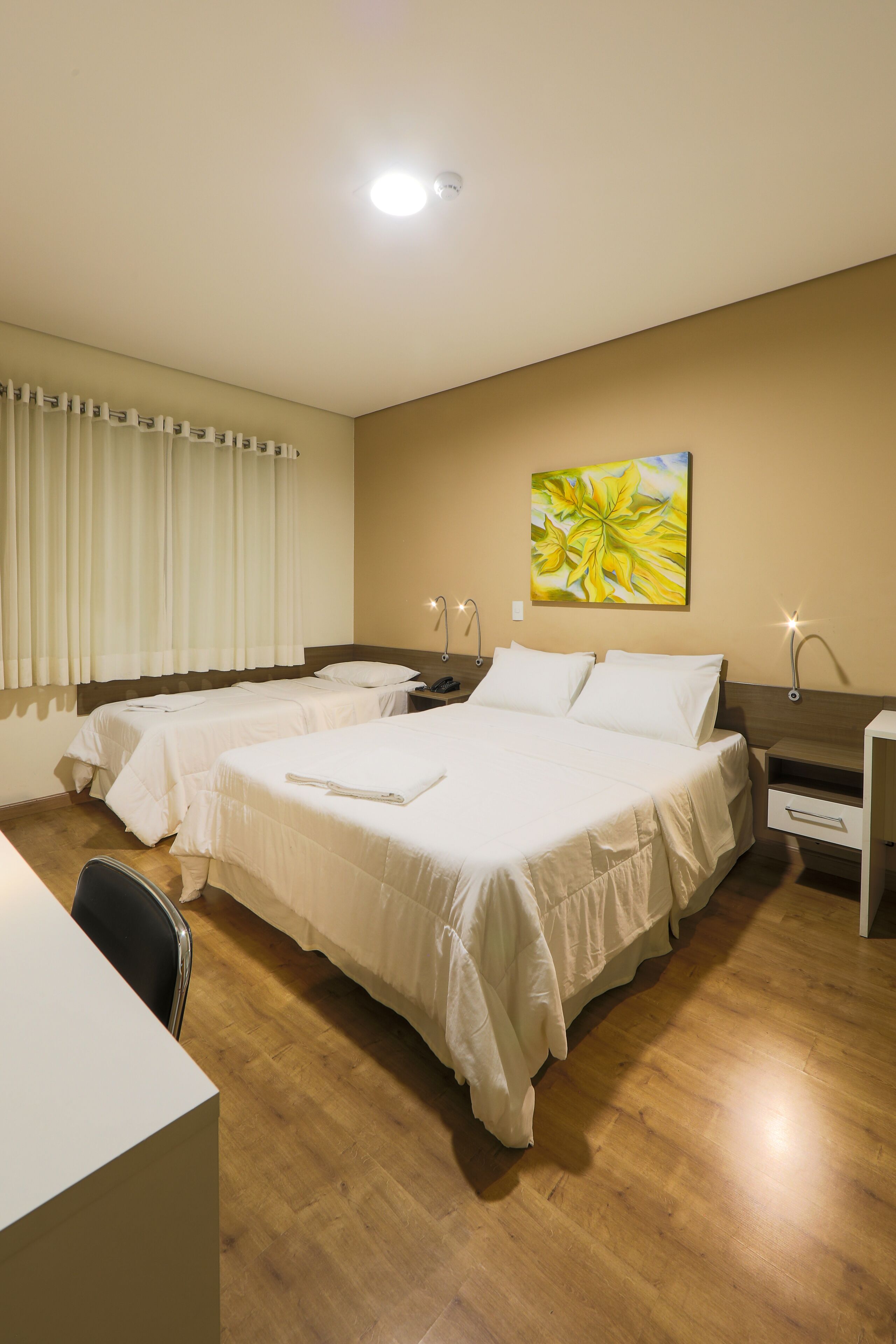 Quarto de hotel com duas camas, uma escrivaninha, uma cadeira e uma mesa de cabeceira com abajur.