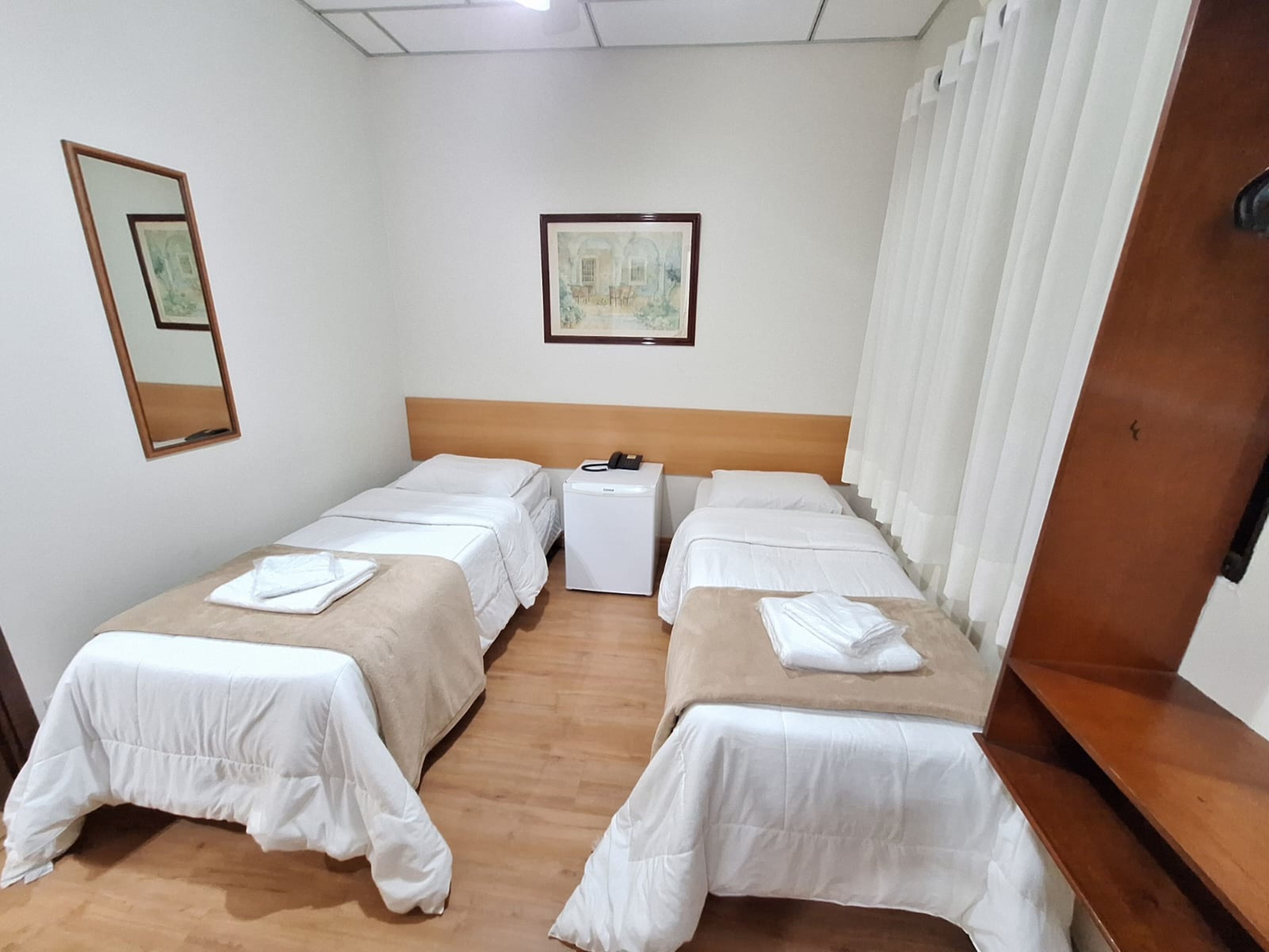 Une chambre d’hôtel avec deux lits simples, une table de chevet, une petite armoire blanche, un miroir et un tableau encadré au mur.
