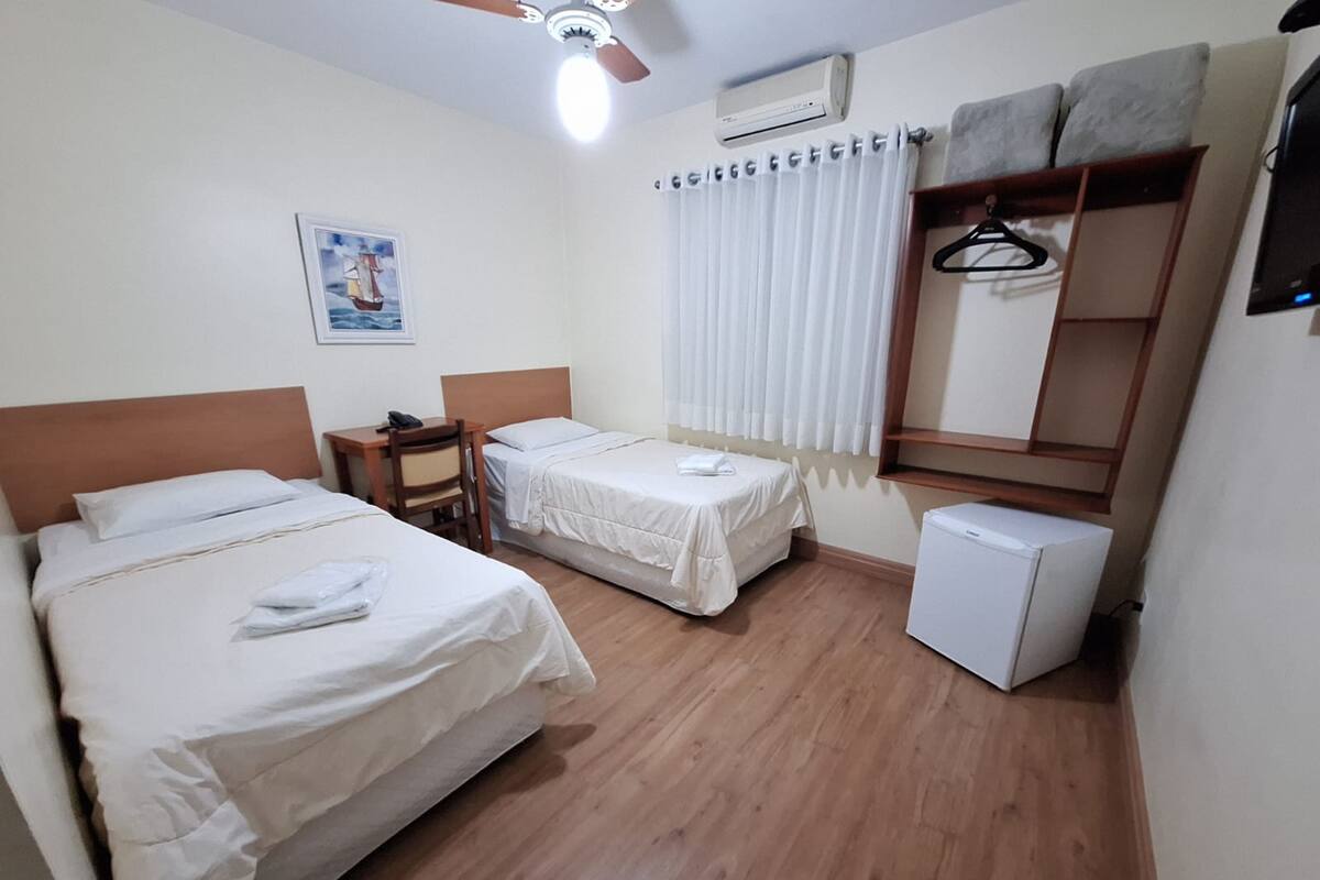 A hotel room with two beds, a desk, a chair, a wardrobe, a heater, and a ceiling fan.