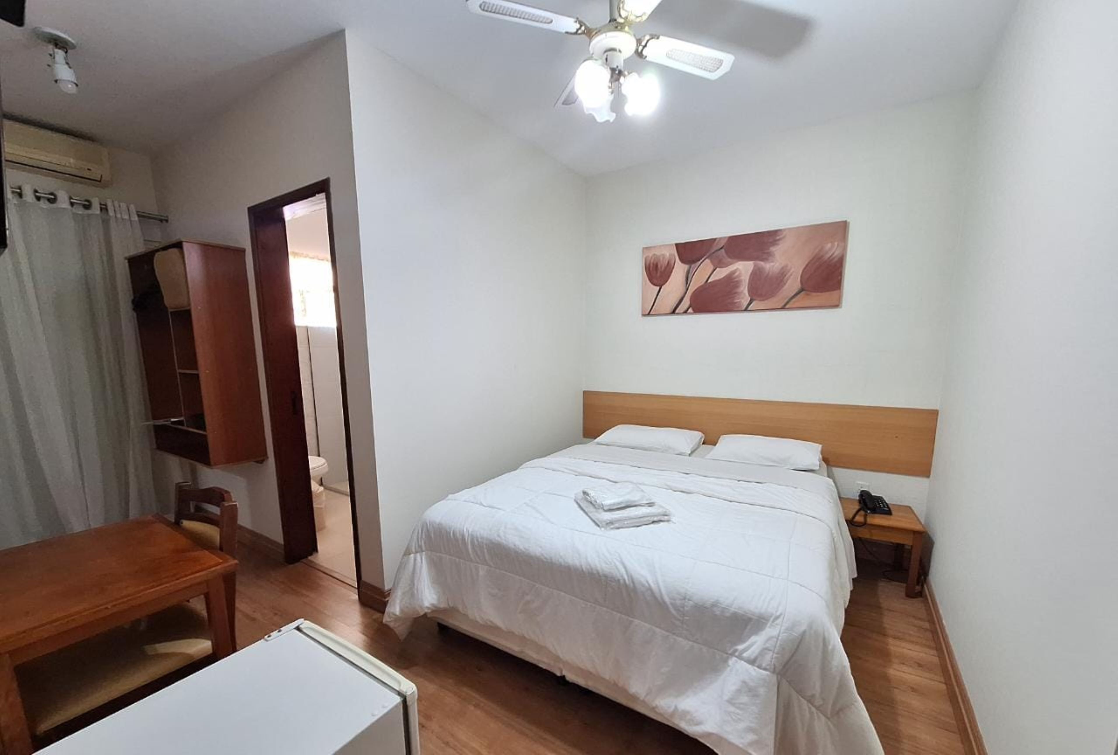 A bedroom with a bed, a wooden headboard, a nightstand with a phone, a ceiling fan, and a painting on the wall.