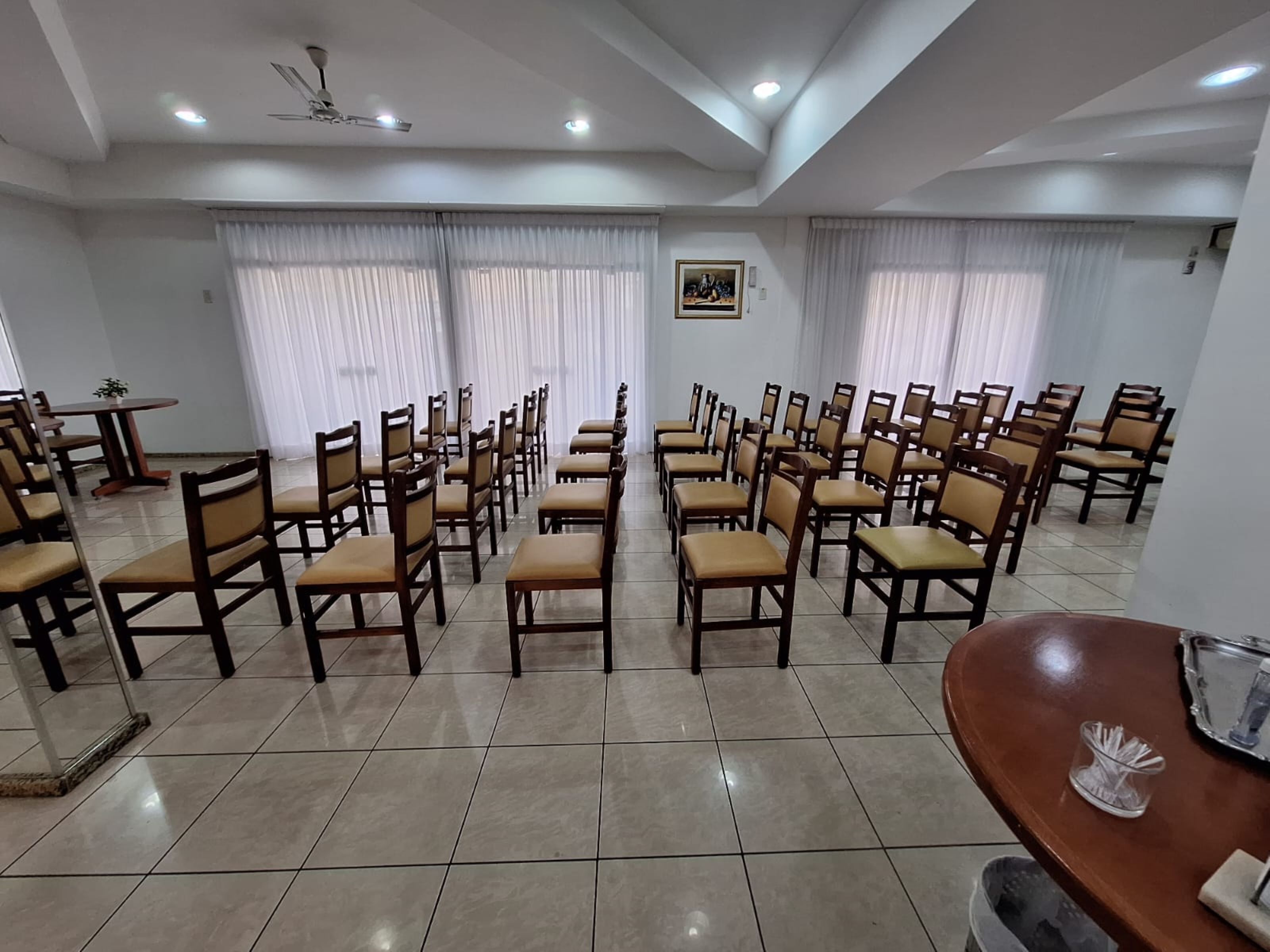 A conference room with rows of wooden chairs and tables, large windows with white curtains, and a ceiling fan.