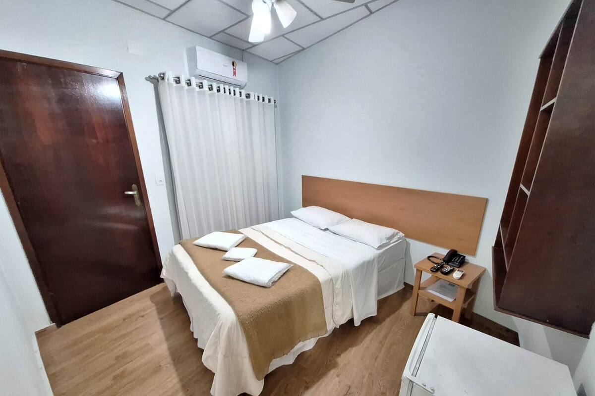 A hotel room with a wooden floor, a bed with white bedding, a wooden nightstand with a phone, and an air conditioning unit on the wall.