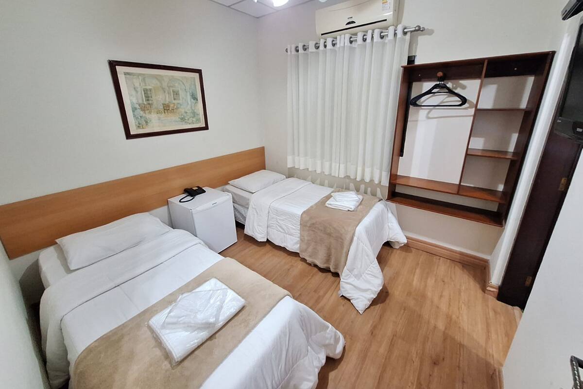 A hotel room with two single beds, a small bedside table with a phone, a ceiling fan, an air conditioning unit, a wardrobe with a mirror, and a framed picture on the wall.