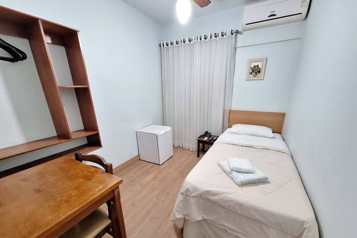 A hotel room with a wooden desk, a bed with white linens, a small bedside table with a phone, and a radiator.