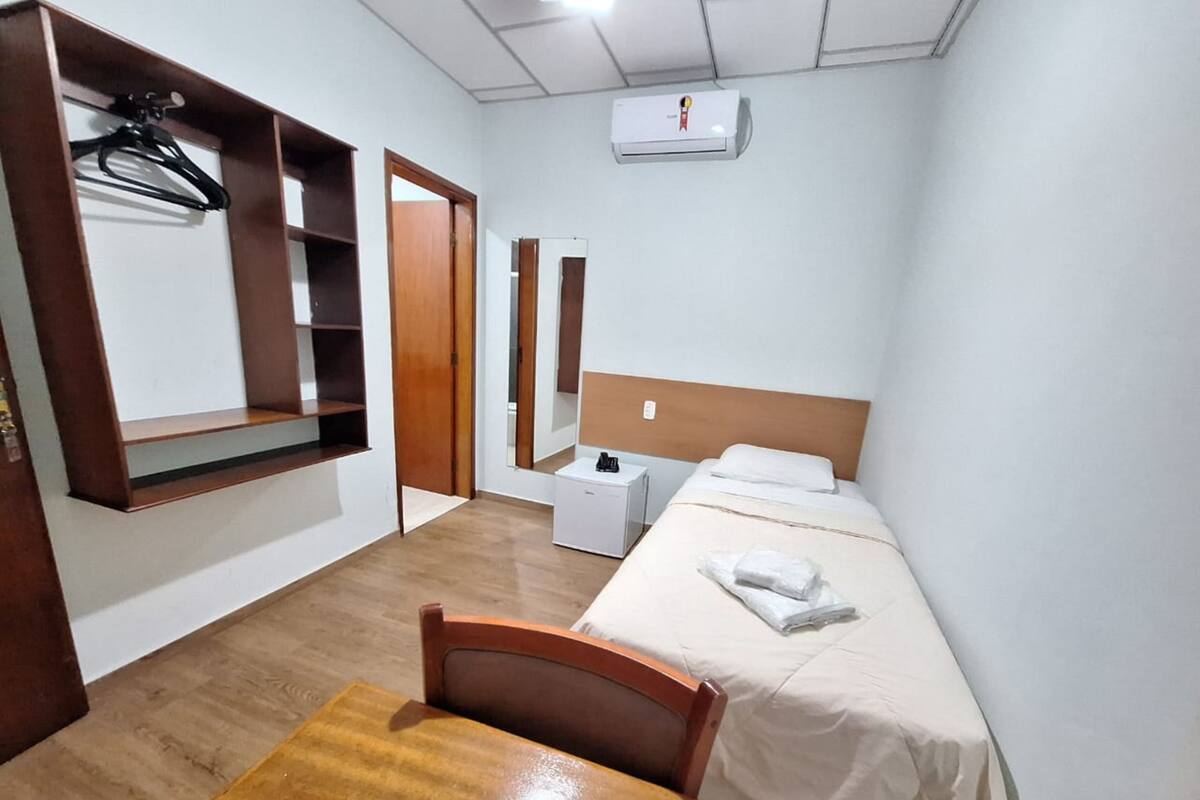 A hotel room with a wooden desk, a bed with white linens, a small bedside table, a mirror, and a wall-mounted air conditioning unit.