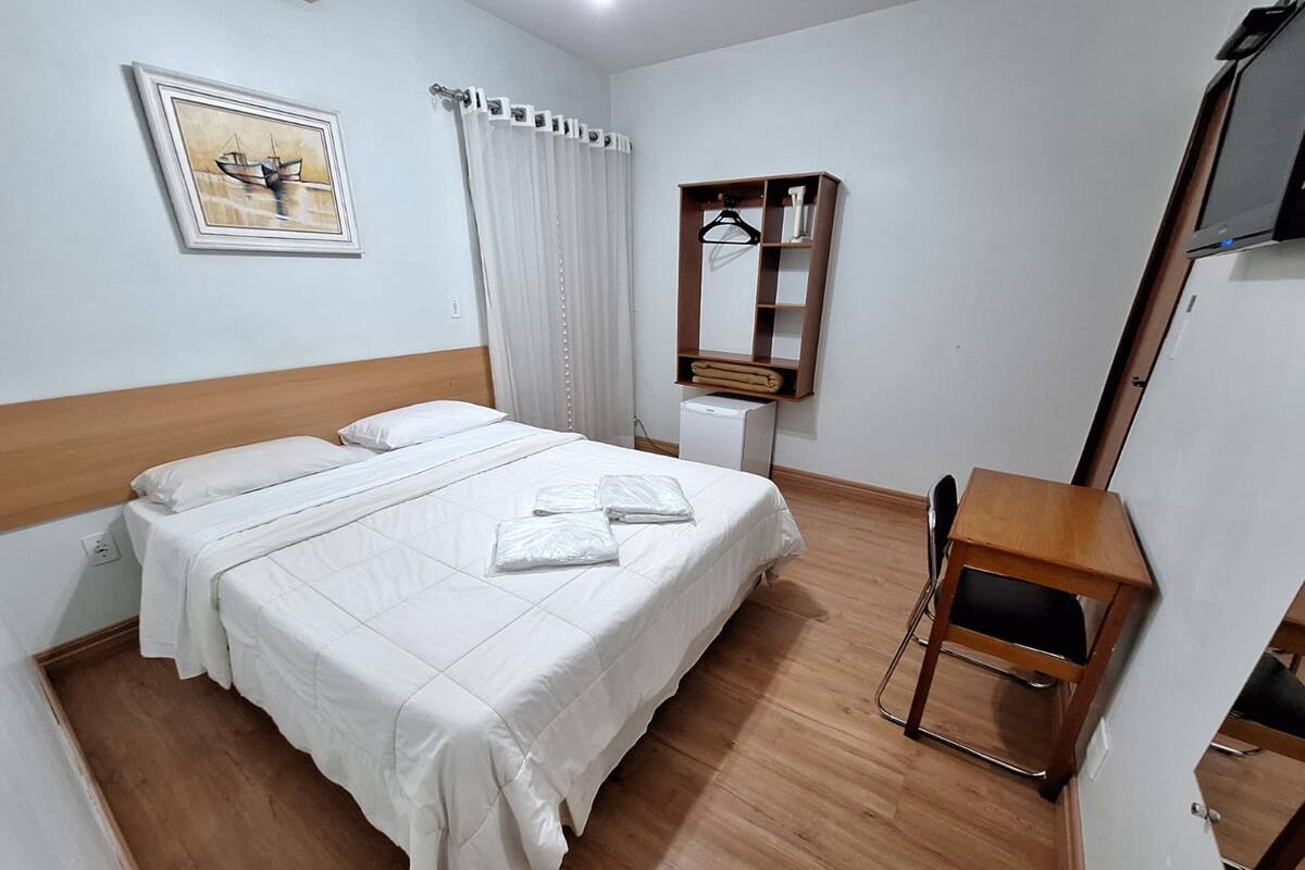 A hotel room with a bed, a desk, a chair, a mirror, a wardrobe, and a painting on the wall.